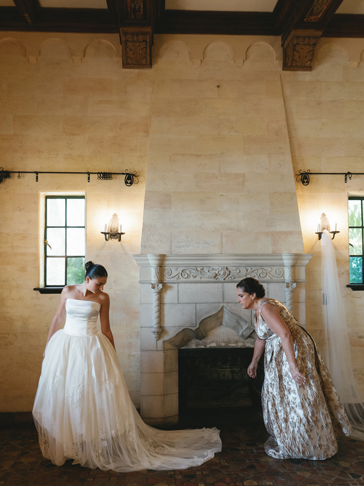A bride in a strapless white wedding gown with a full tulle skirt stands to the left, looking down. An older woman in an ornate gold dress bends down near a fireplace, adjusting her dress. The room has a high ceiling, stone walls, two small windows, and decorative wall-mounted light fixtures.