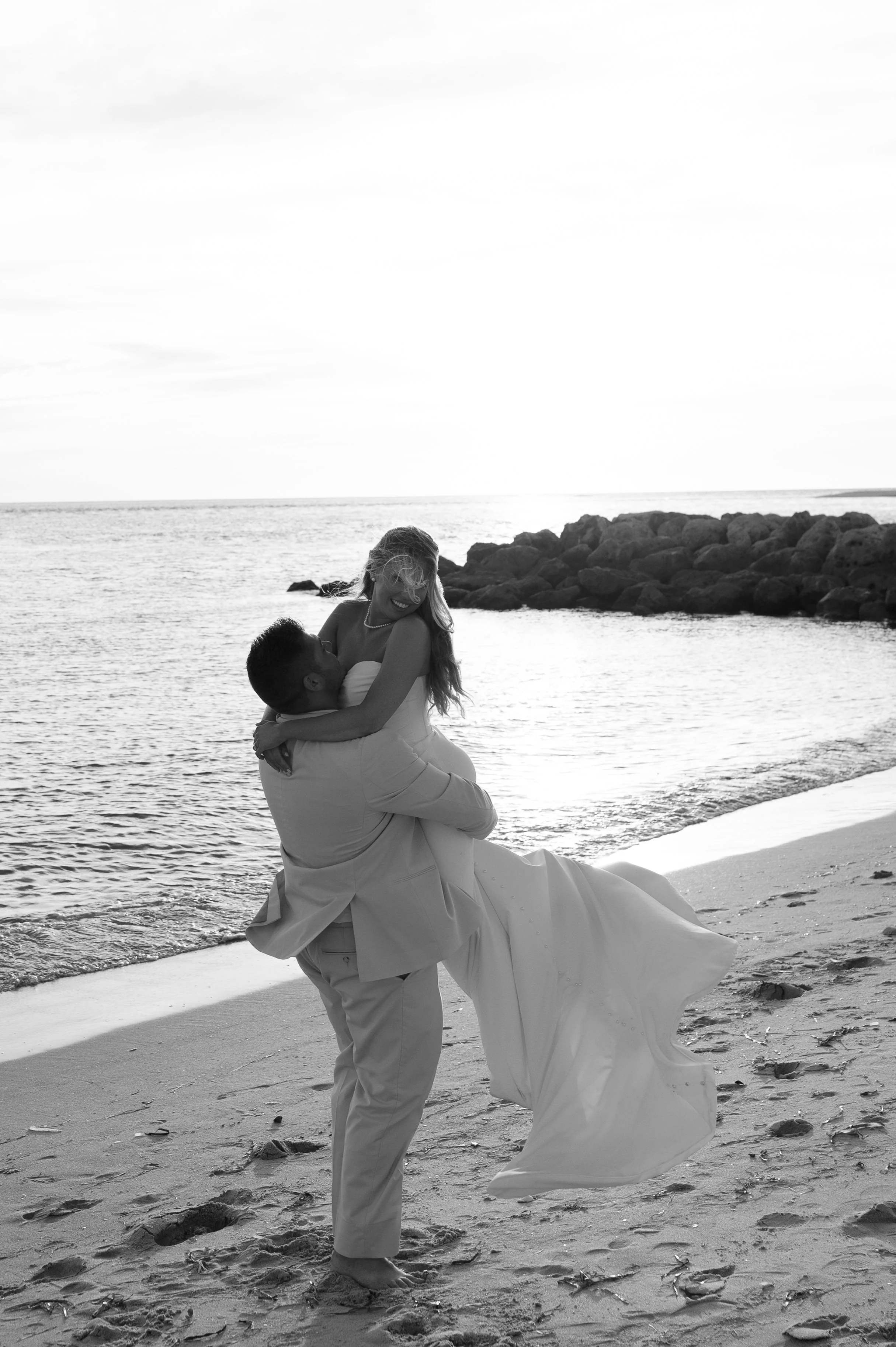 A black and white photo of a man lifting a woman in a wedding dress on a beach with rocks, water, and cloudy sky in the background.