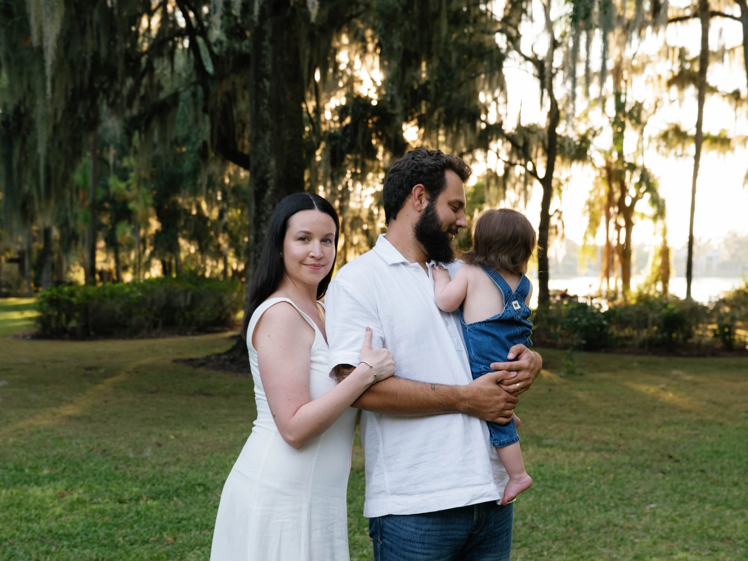 A woman in a white dress gently holds her partner, who is holding a small child, as they stand on a grassy area during sunset among tall trees draped with moss.