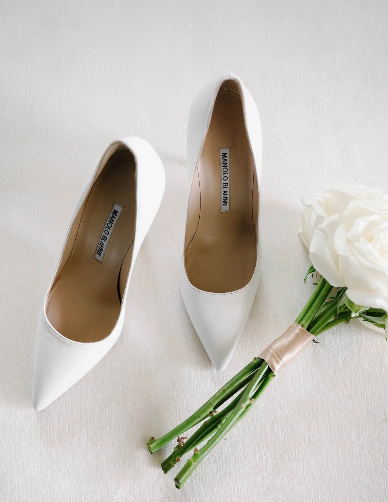 A pair of white pointed-toe high heels with tan insoles, a bouquet of white flowers wrapped in beige ribbon, laid on a white surface.