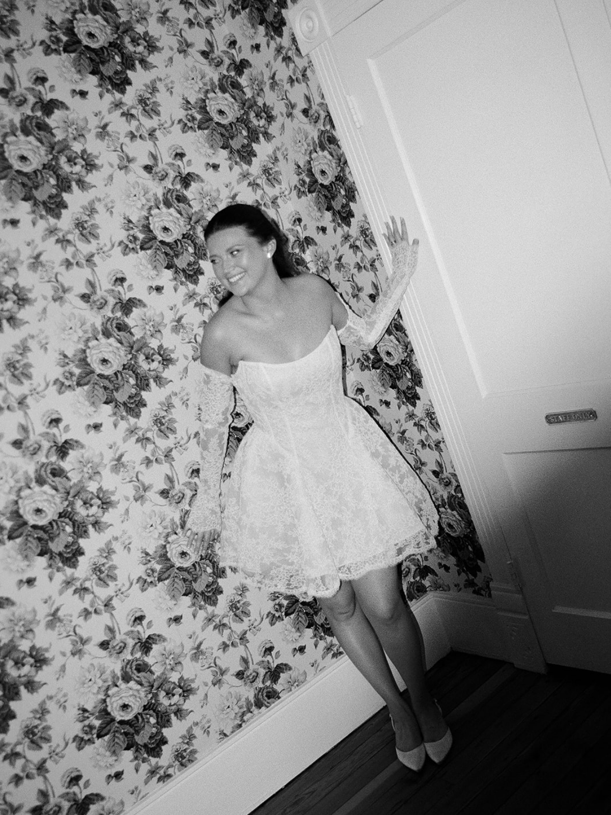 A woman in a wedding dress standing next to a floral wall, smiling and touching a door.