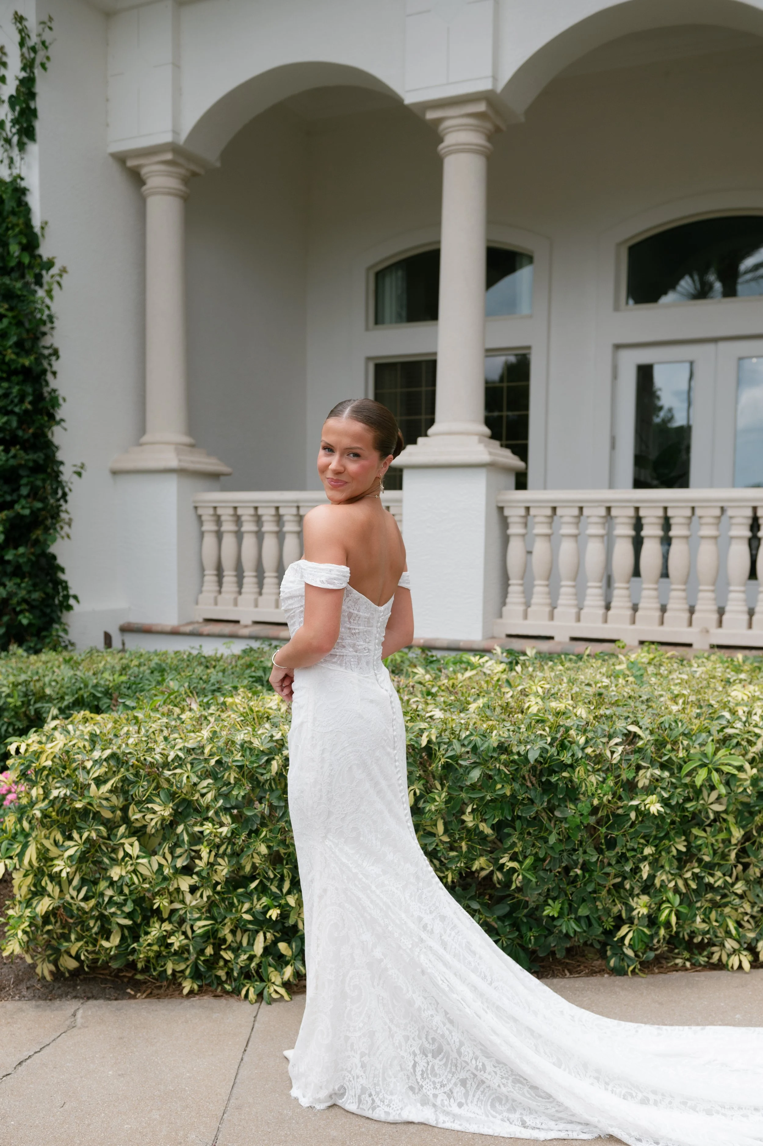 A woman in a white wedding gown standing outside in front of a white house with arched columns and large windows. She is smiling and looking over her shoulder.
