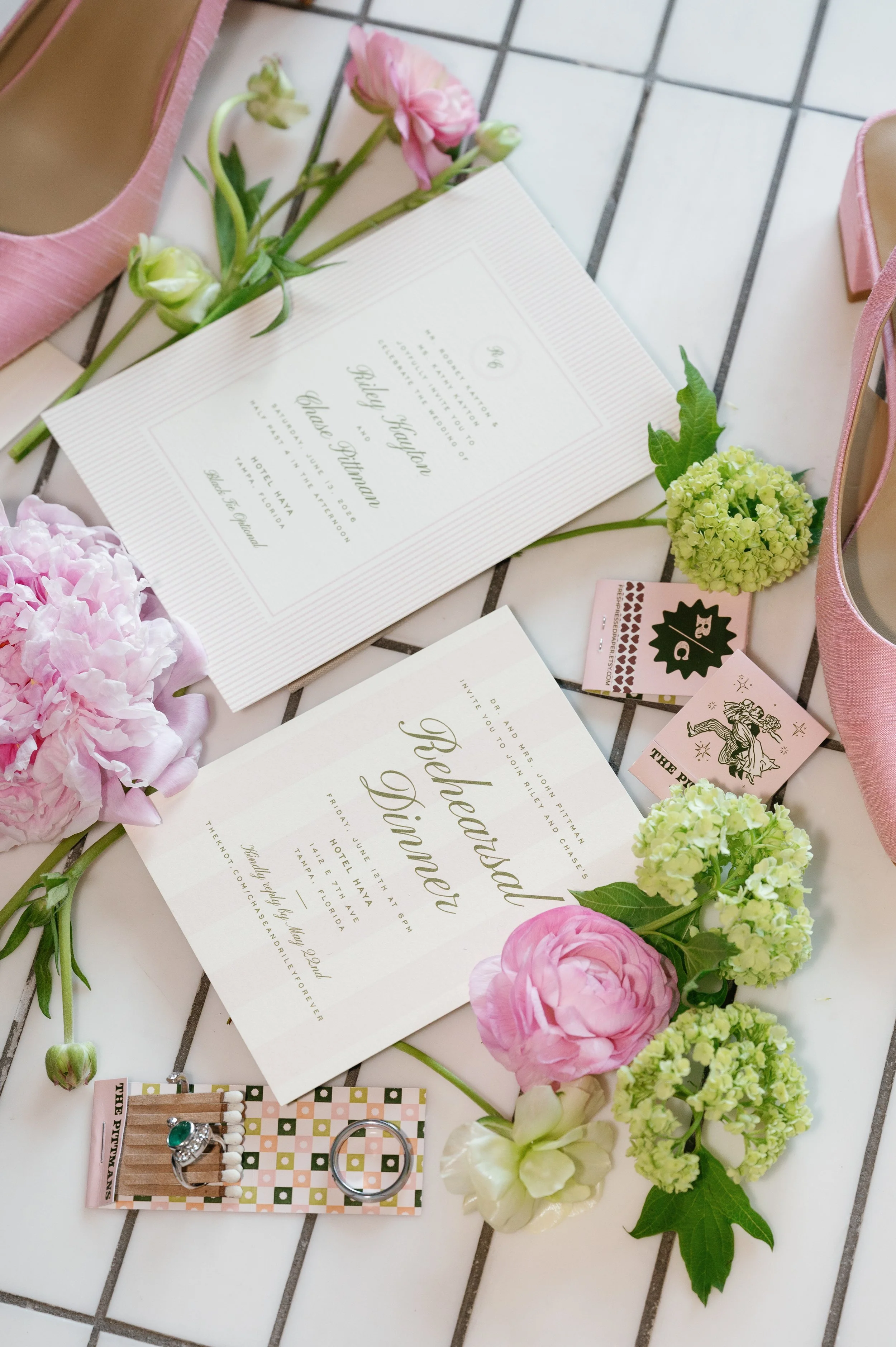 Wedding invitation with pink and green flowers, pink shoes, matchbox, and jewelry placed on a tiled floor.