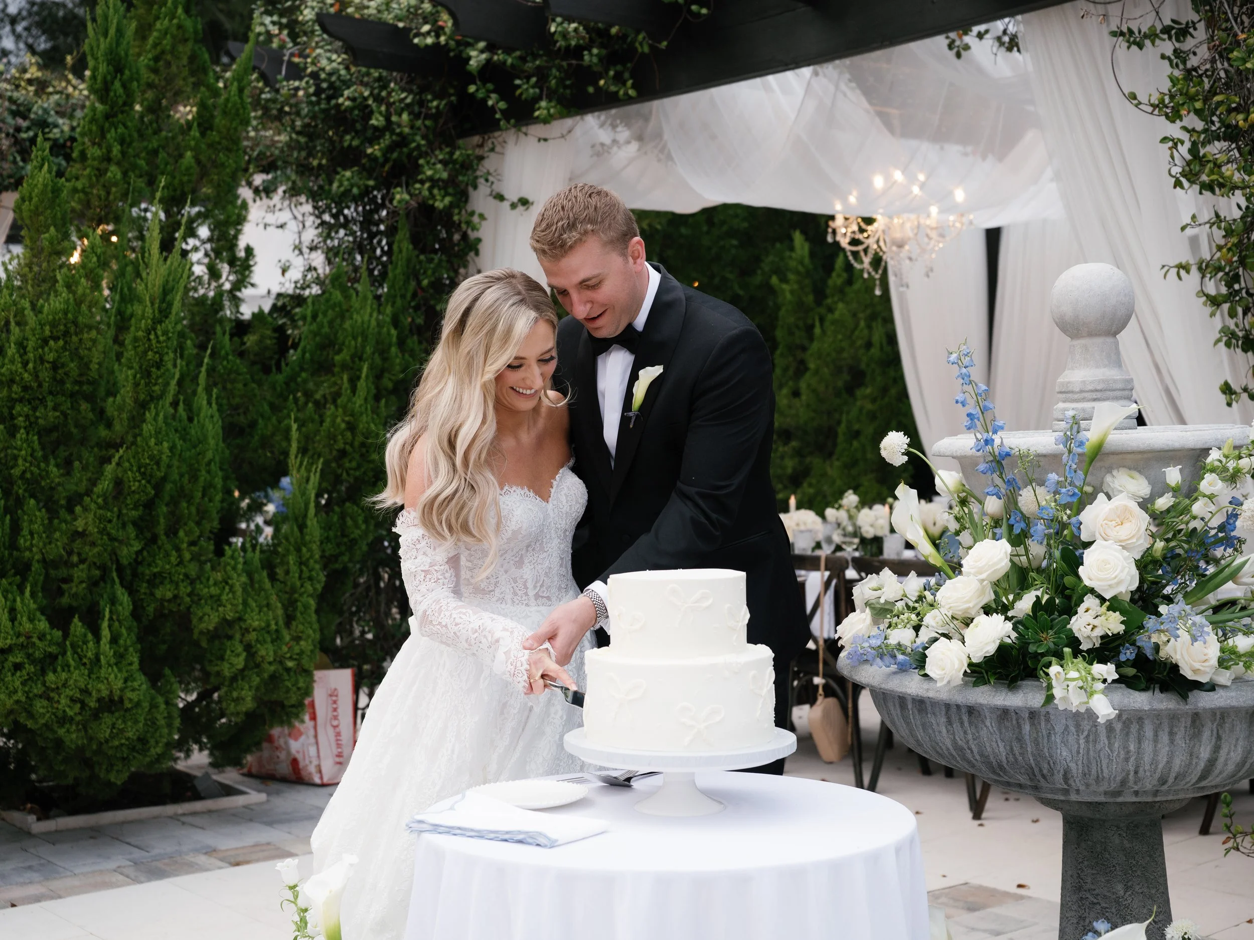 A bride and groom cutting a wedding cake at a reception outdoors, with greenery and floral arrangements in the background, a large fountain with flowers in the foreground, and a chandelier hanging from a decorated canopy.