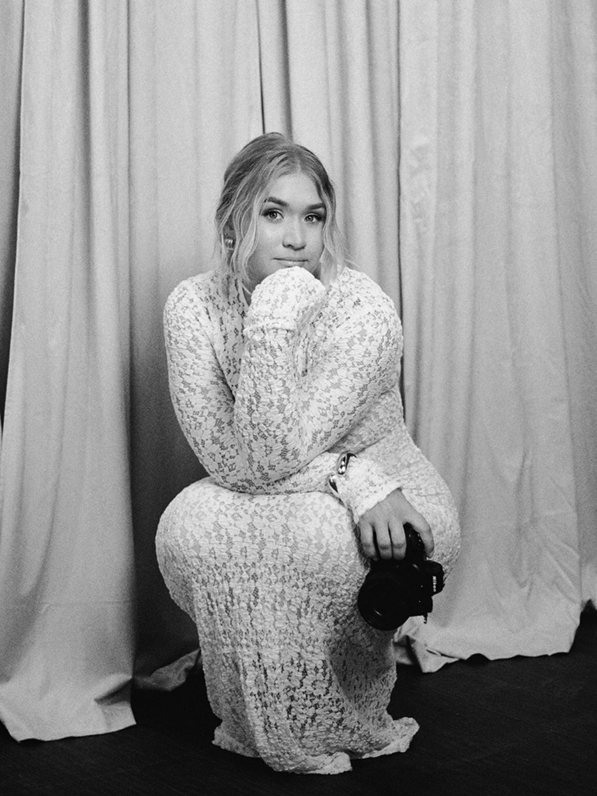 Black and white photo of a young woman in a lace dress, sitting on a chair, holding a camera, with a curtain background.