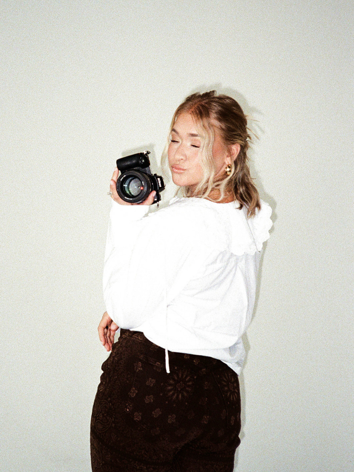 A woman with blonde hair holding a camera and making a kiss face, wearing a white hoodie and brown patterned pants, standing against a plain light-colored background.