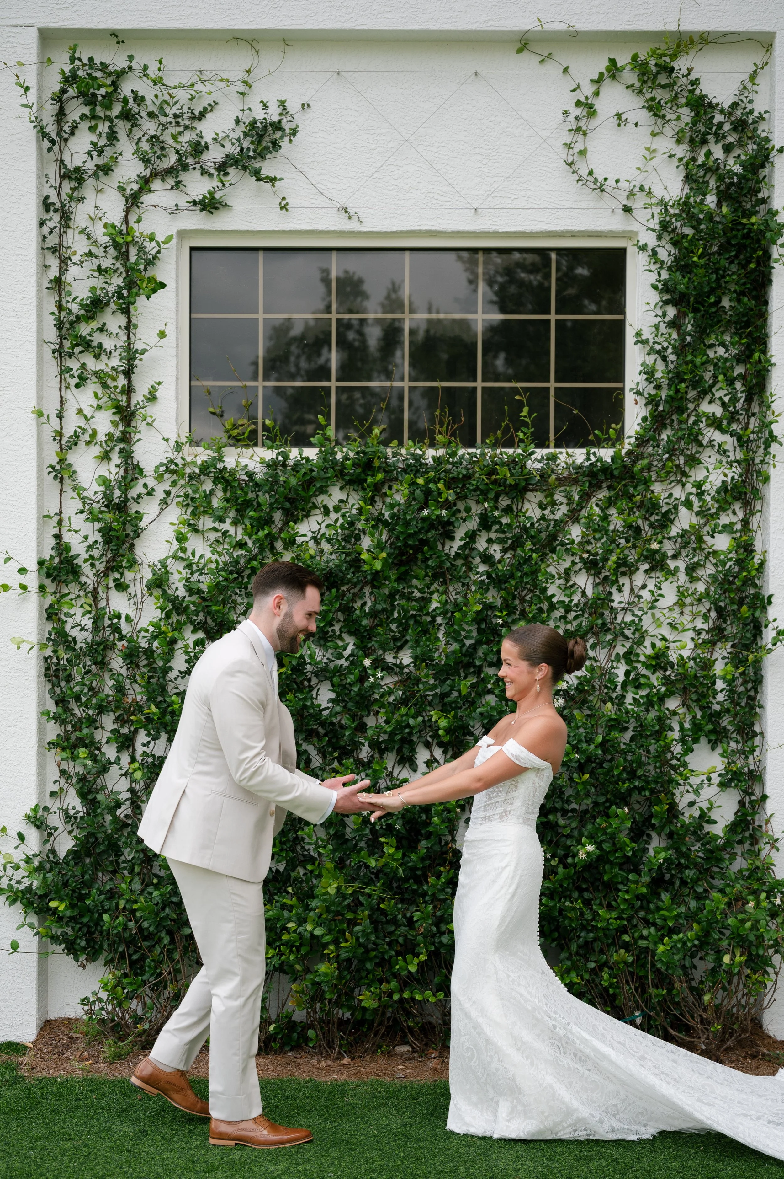 A bride and groom exchanging vows outdoors in front of a white wall with greenery. The groom is wearing a light-colored suit with brown shoes, and the bride is in a white lace wedding gown with a train. They are holding hands and smiling at each othe
