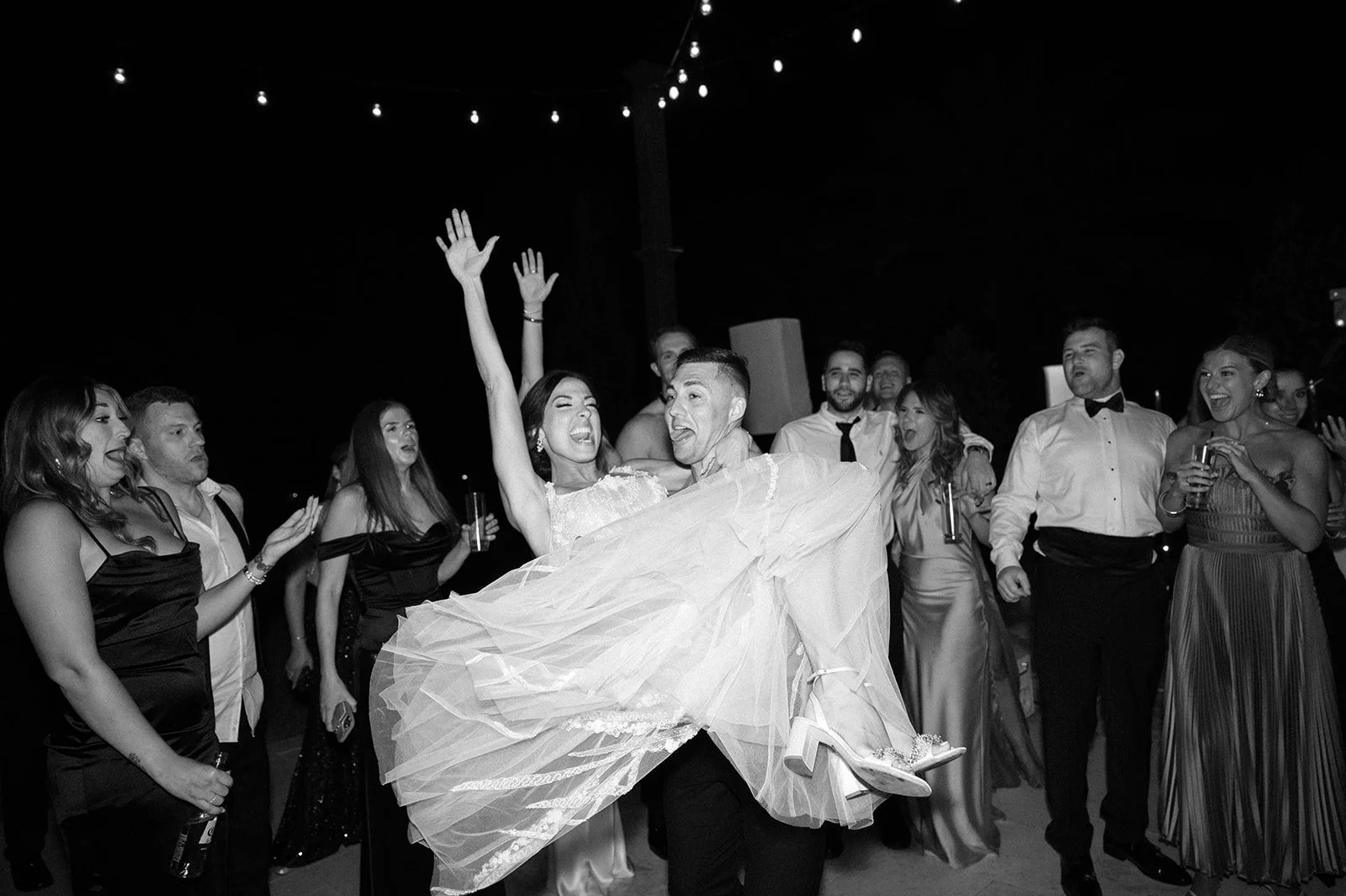 A bride in a wedding dress is lifted by a groom at a wedding reception, surrounded by guests celebrating and dancing.