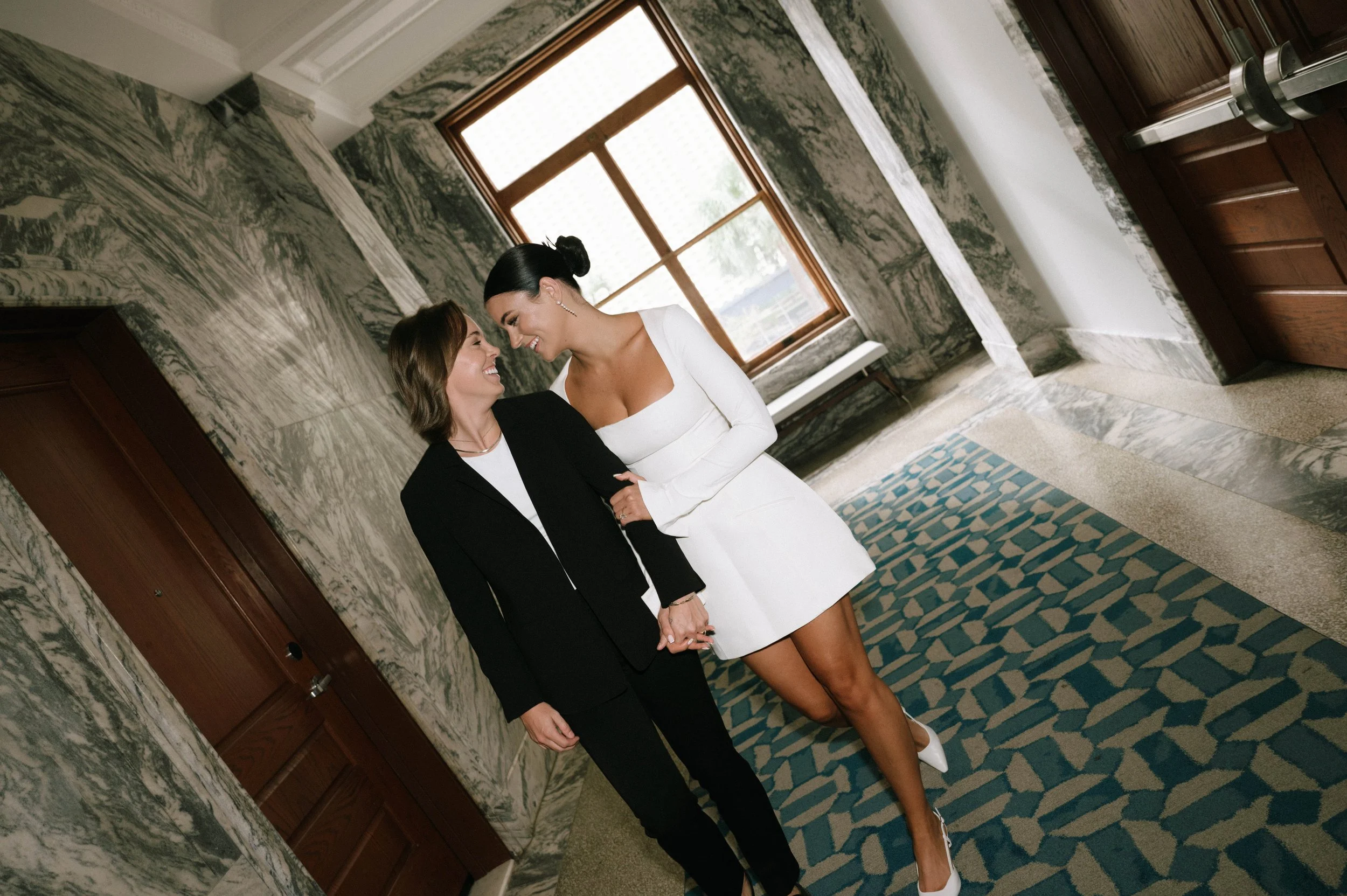 Two women holding hands and smiling at each other in a hallway with marble walls and a window.