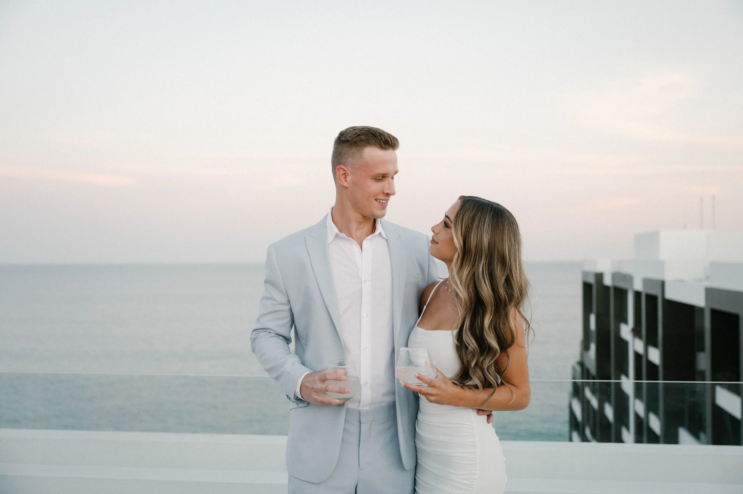 A couple dressed in white attire standing on a deck by the water, holding glasses of wine, with a city skyline in the background.