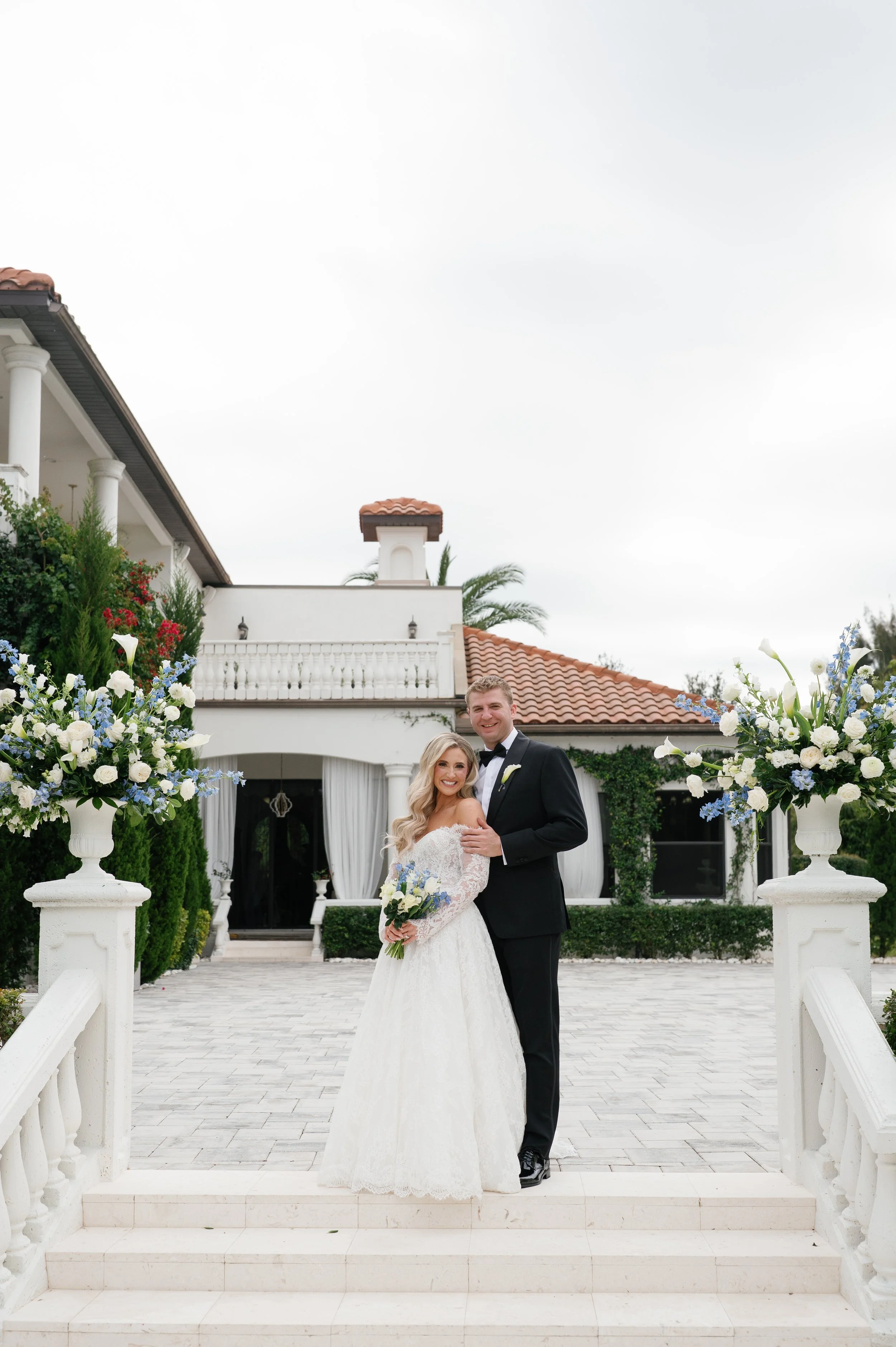 A bride and groom standing on white stairs in front of a house with white columns and a red-tiled roof, surrounded by floral arrangements, on a cloudy day.