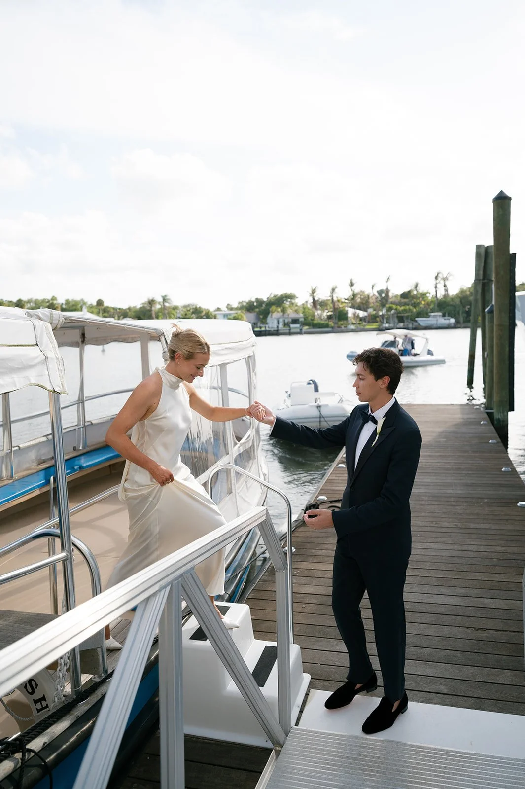 A bride in a white dress is stepping onto a boat, assisted by a groom in a black tuxedo, at a dock by the water on a sunny day.