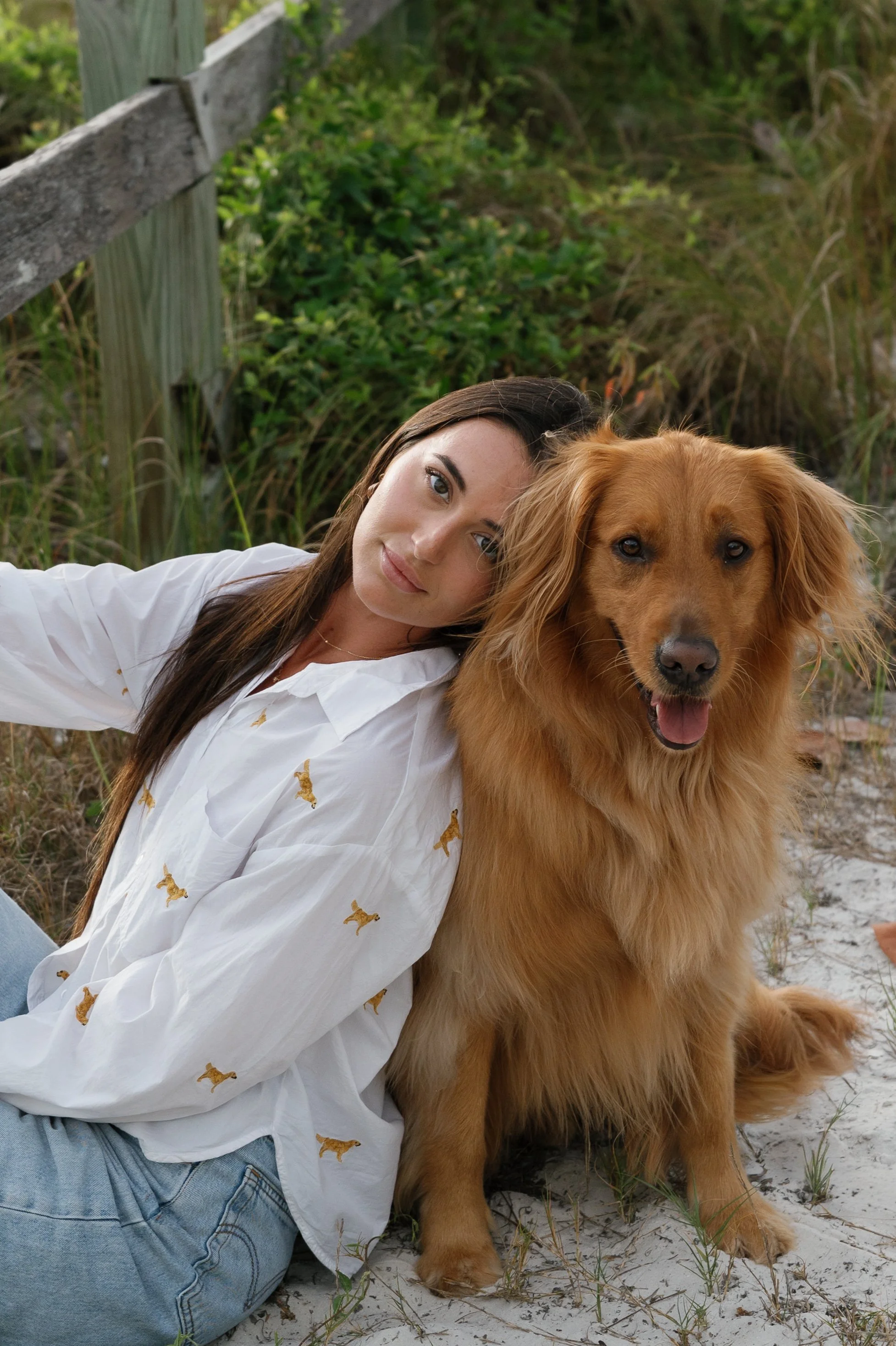A woman with long brown hair lying on sandy ground next to a golden retriever dog, both looking at the camera. The woman is wearing a white shirt with small dog prints and jeans. The background includes green shrubs, tall grass, and a weathered woode