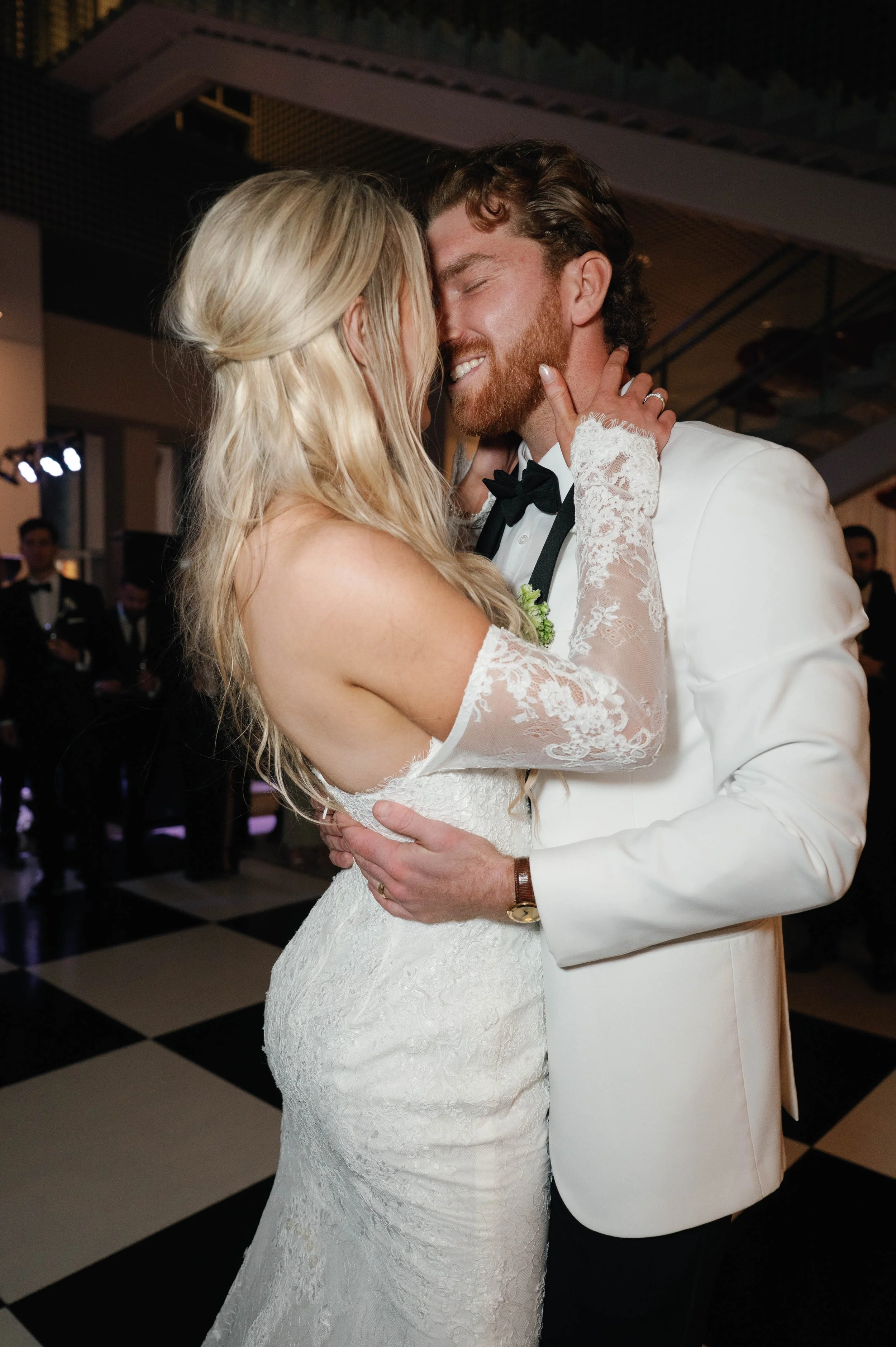 A bride and groom are dancing closely during their wedding reception, with the bride wearing a lace wedding gown and the groom in a white tuxedo with a black bowtie, in a ballroom with black and white checkered floor.