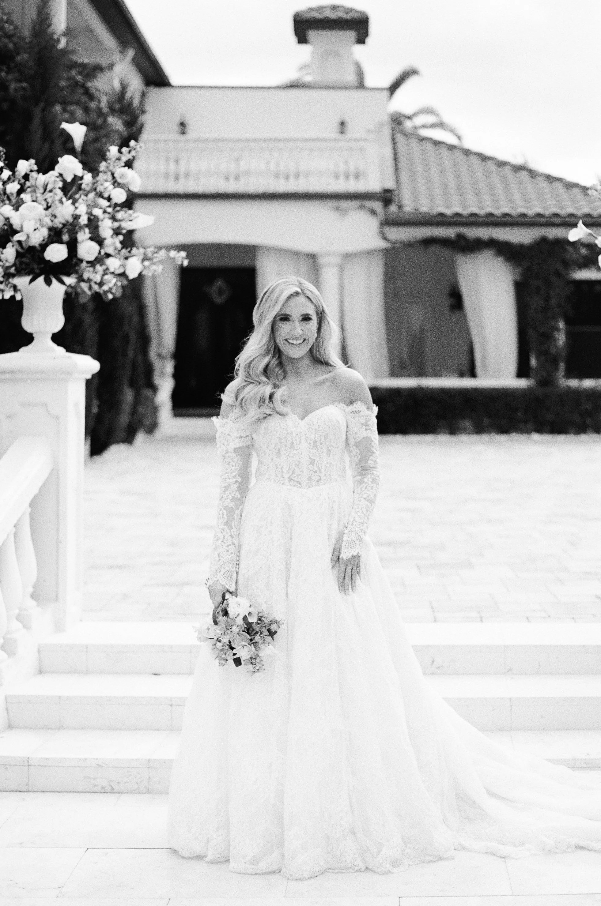 Black and white photo of a smiling woman in a wedding gown standing on steps outside a house, holding a bouquet of flowers.