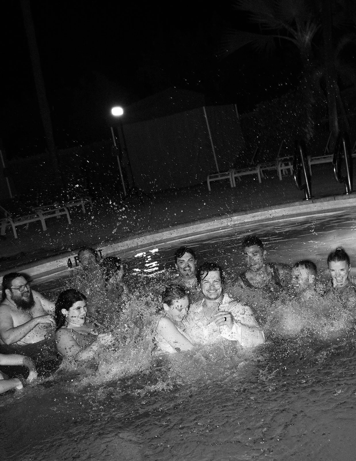People enjoying a pool at night, smiling and splashing water, with pool chairs and a fence visible in the background.