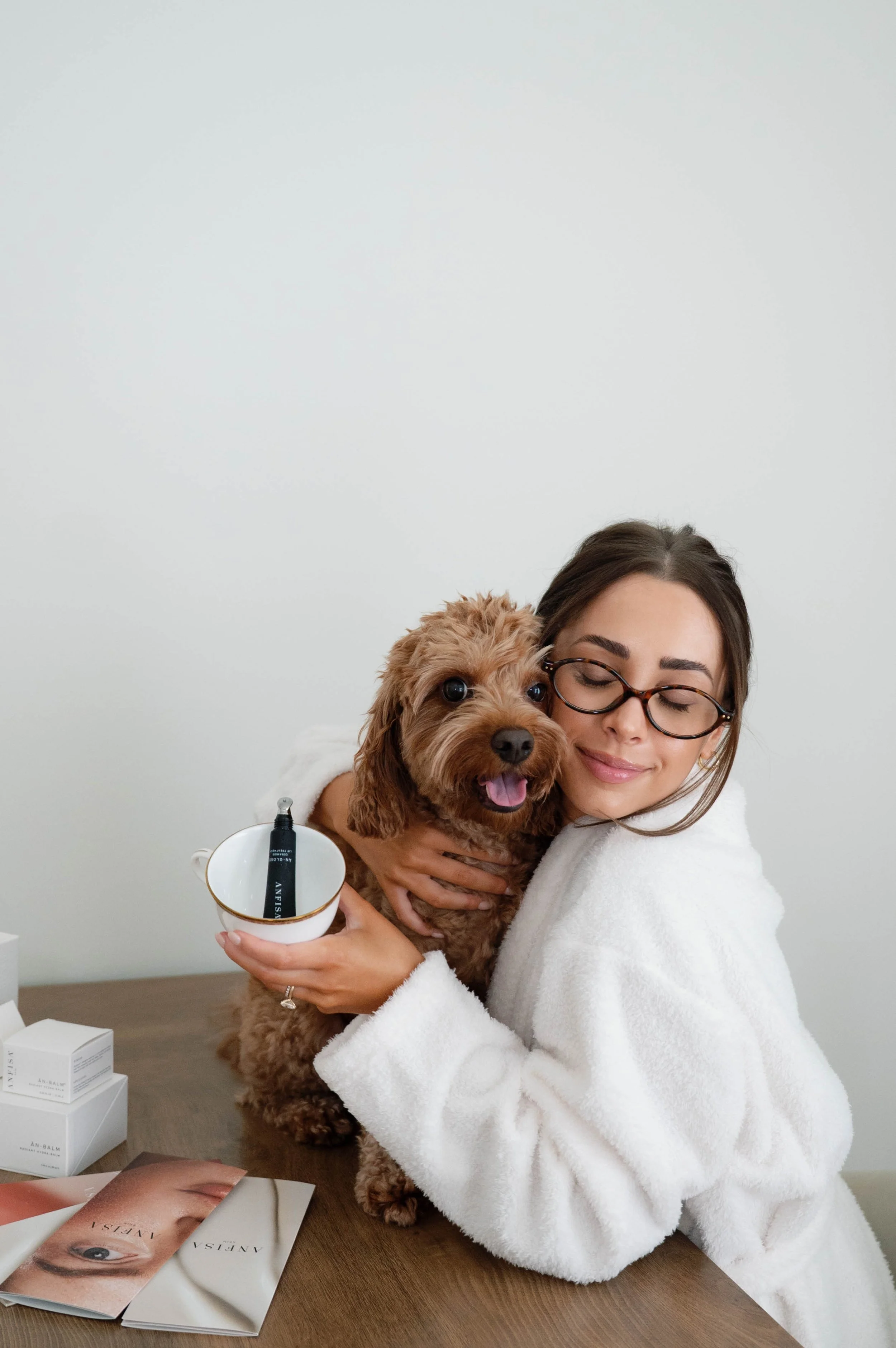 A woman wearing glasses and a white robe hugs a small, curly-haired brown dog while holding a white mug with skincare products inside. The table in front has beauty magazines and skincare boxes.