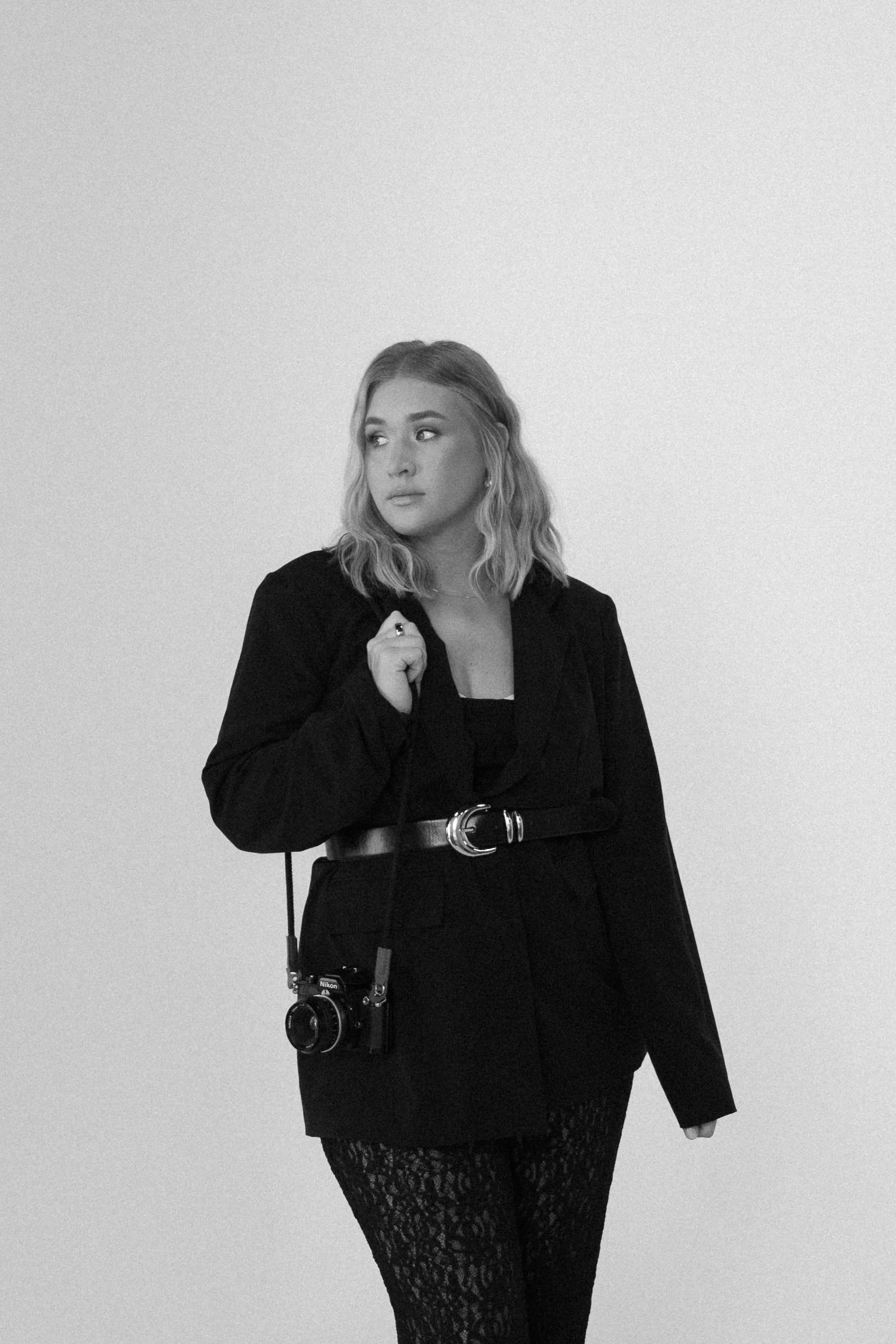 Black and white photo of a woman with wavy hair, wearing a jacket and lace leggings, holding a camera with a strap around her neck, standing against a plain wall.