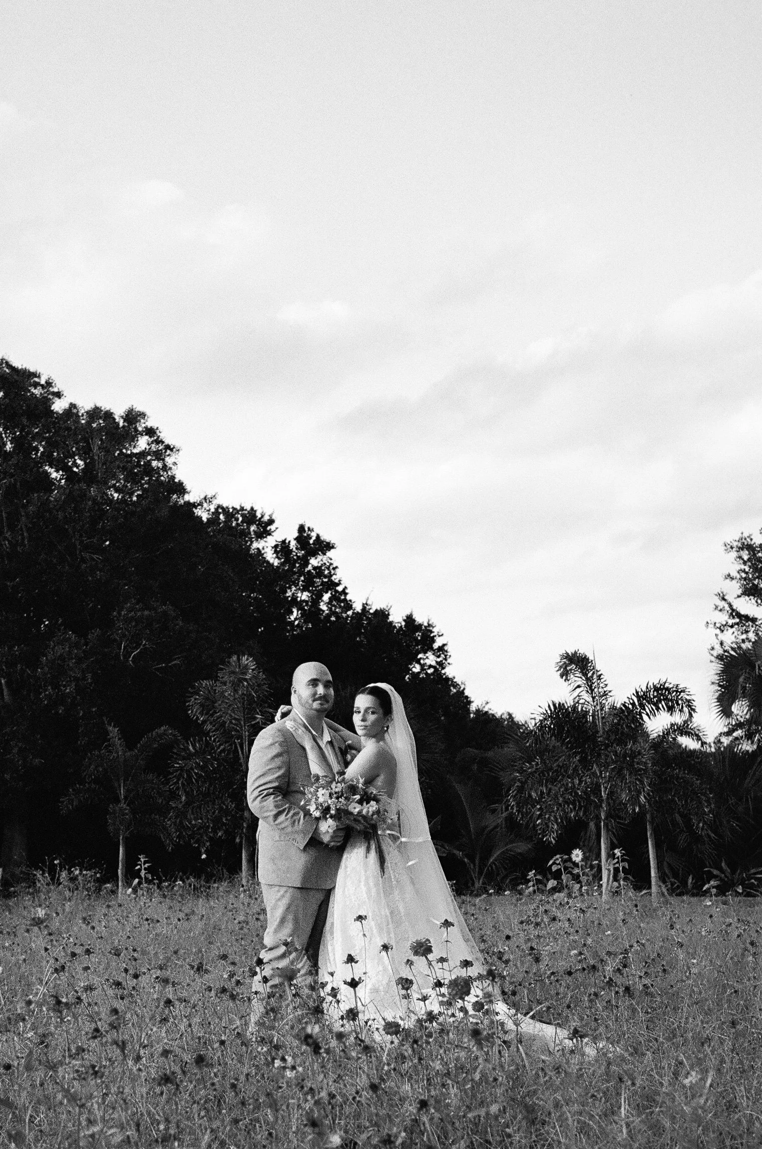 A black and white photo of a bride and groom standing in a field of wildflowers with trees in the background, under an overcast sky.