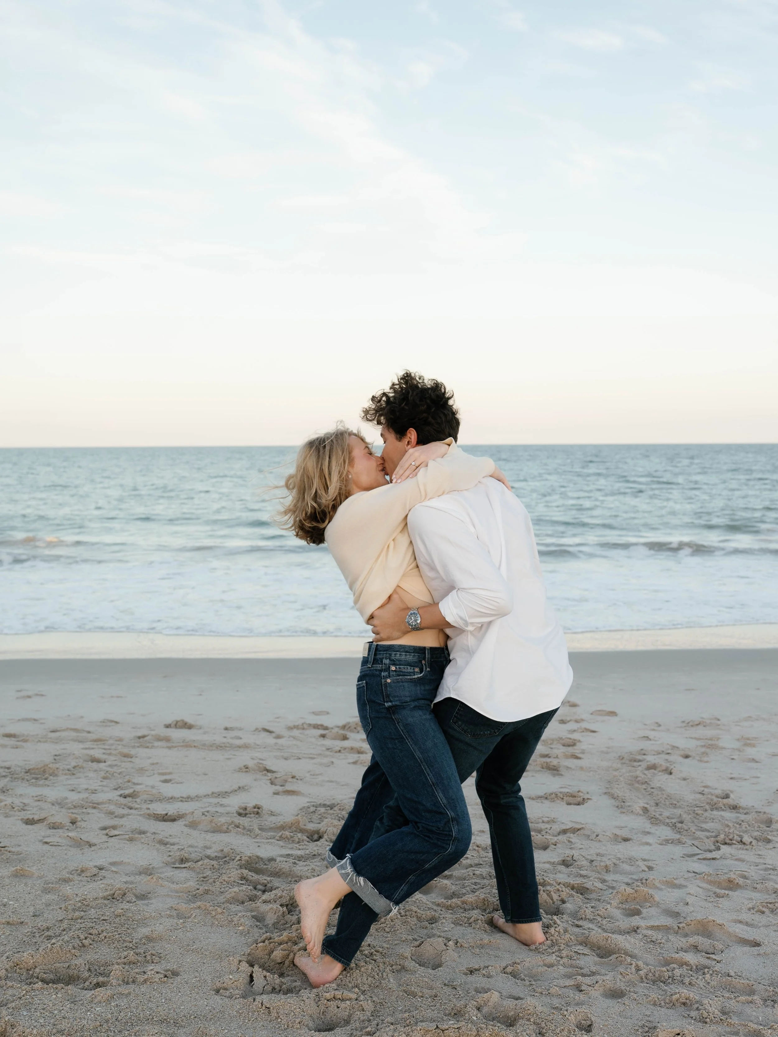 A couple hugging and kissing on a sandy beach near the ocean, with the sky and water in the background.