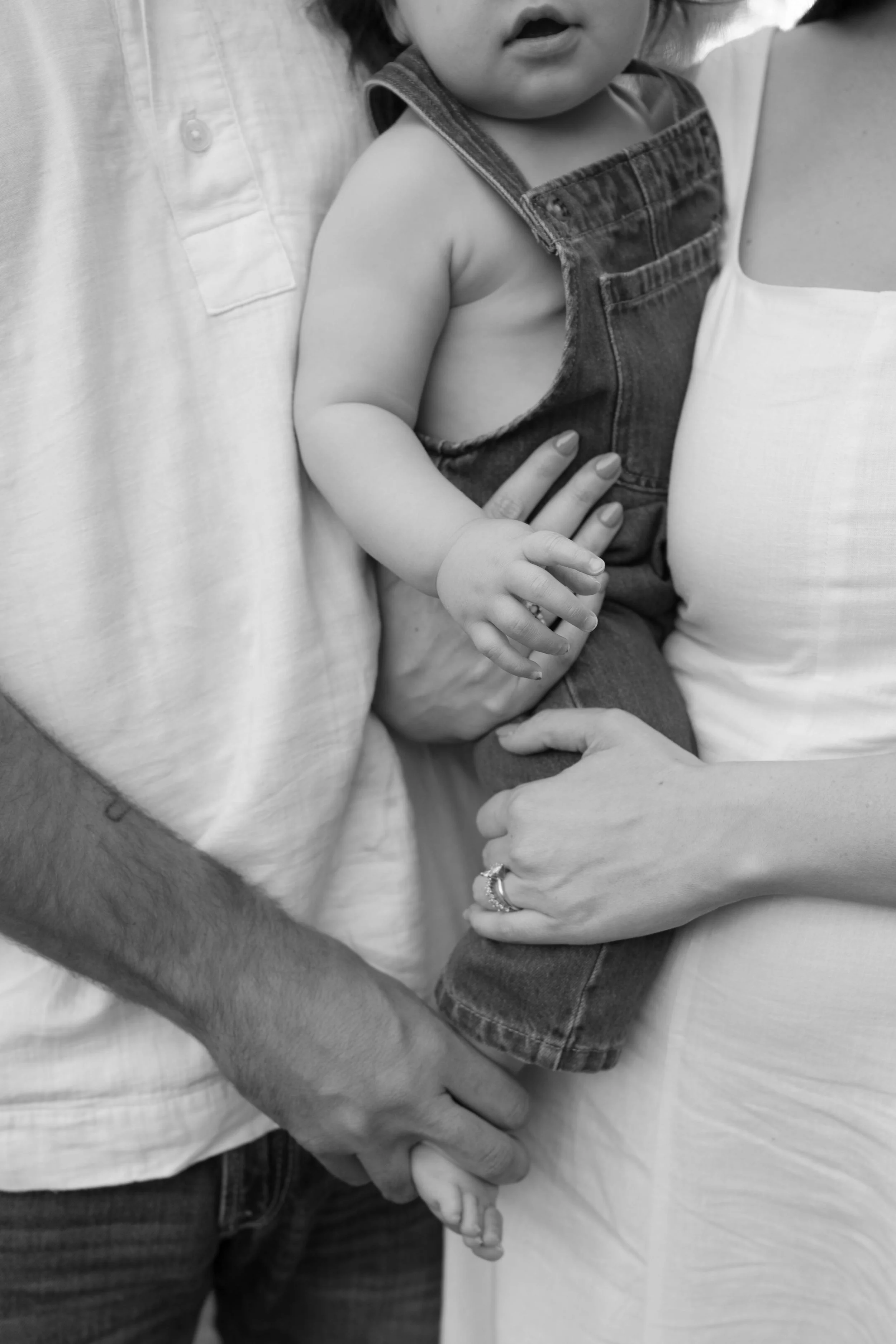 A close-up black and white photograph of a young child being held by two adults, with their arms intertwined. The child is wearing a sleeveless denim top and has their hand resting on an adult's arm.