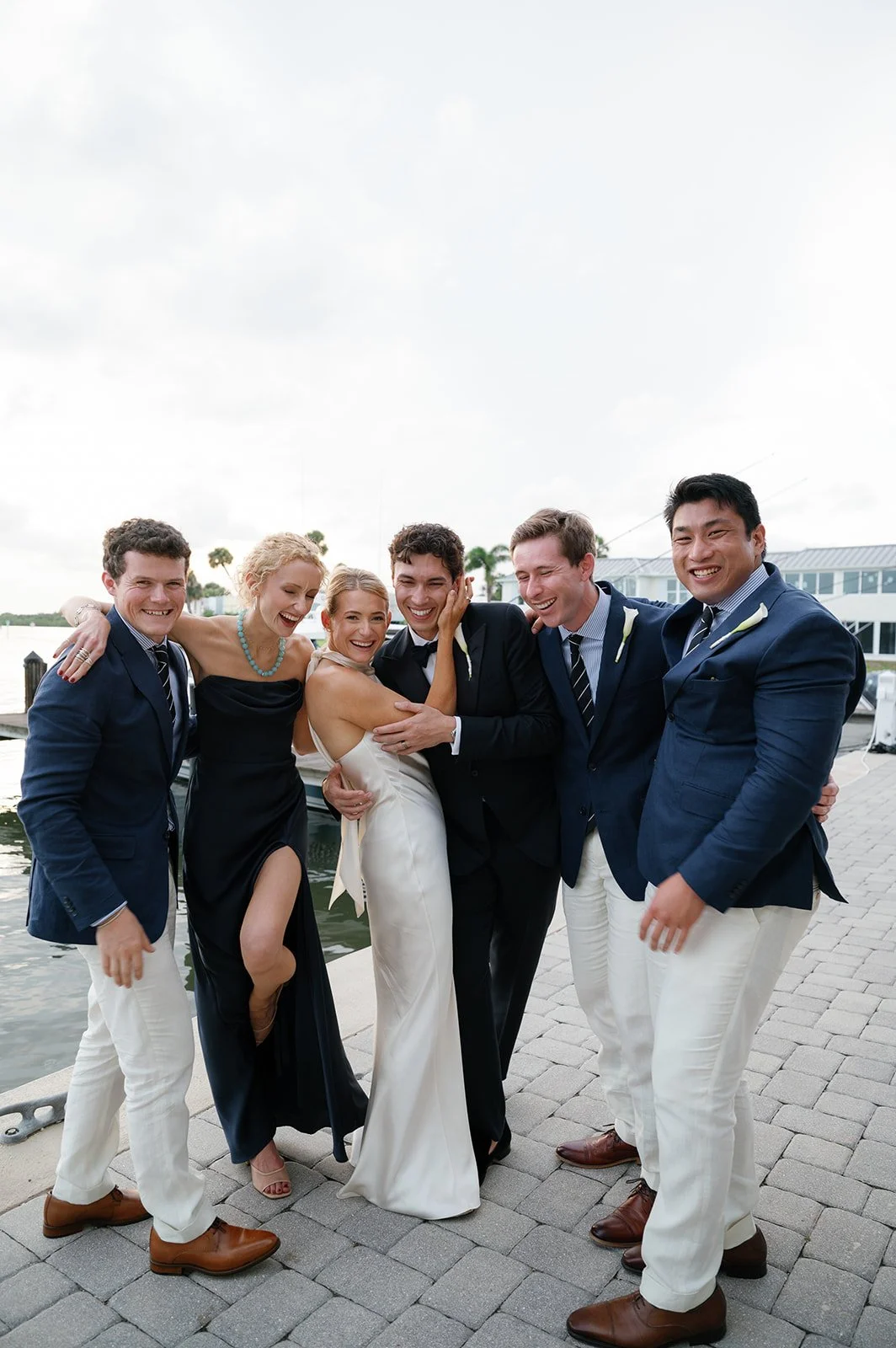 A group of six friends, three men and three women, in formal attire, standing on a waterfront dock, smiling and laughing together.