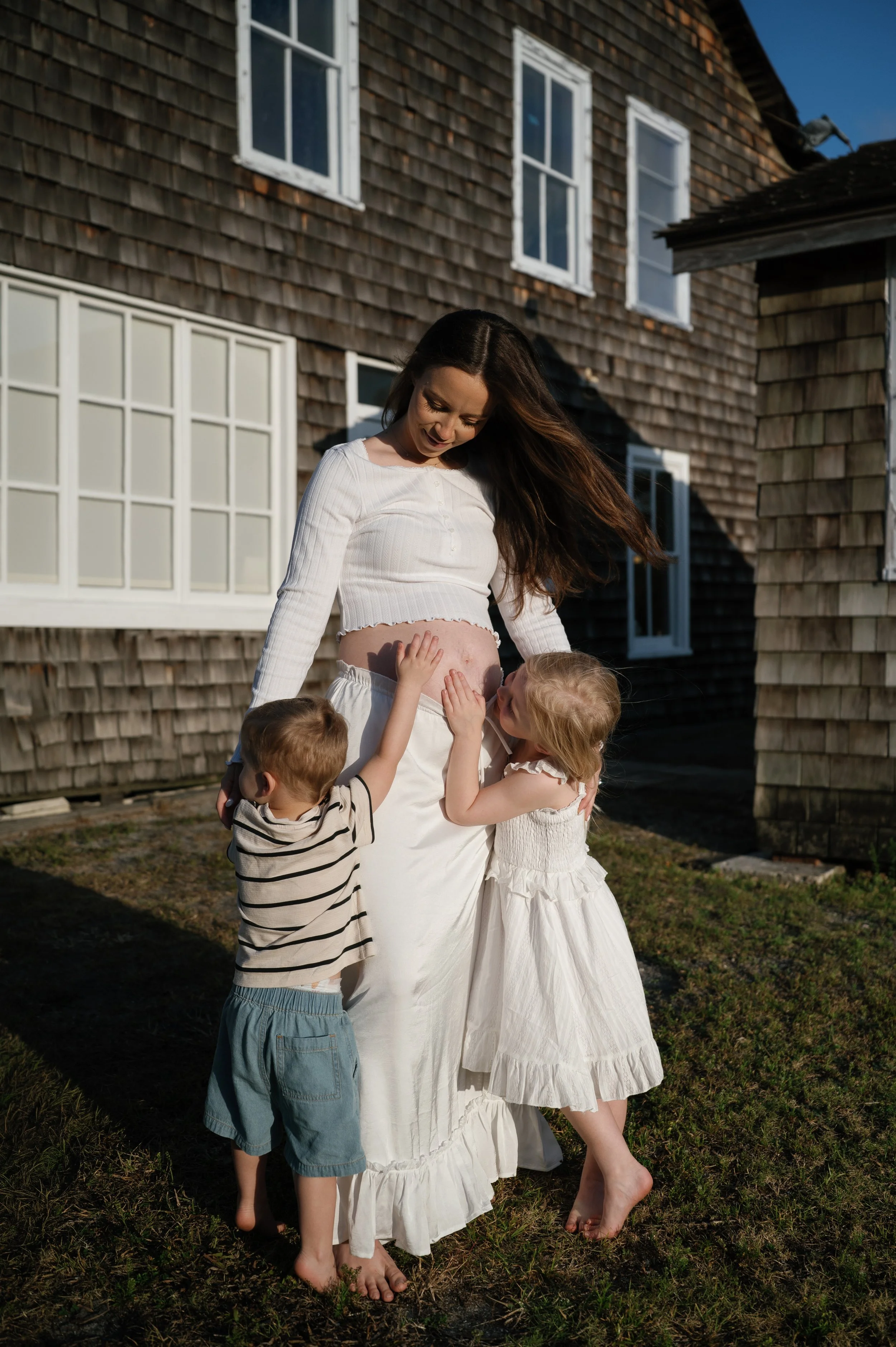 A woman with long dark hair wearing a white crop top and long white skirt is surrounded by four children, two boys and two girls, who are touching her pregnant belly. They are outdoors in front of a weathered wooden house with multiple windows, and t