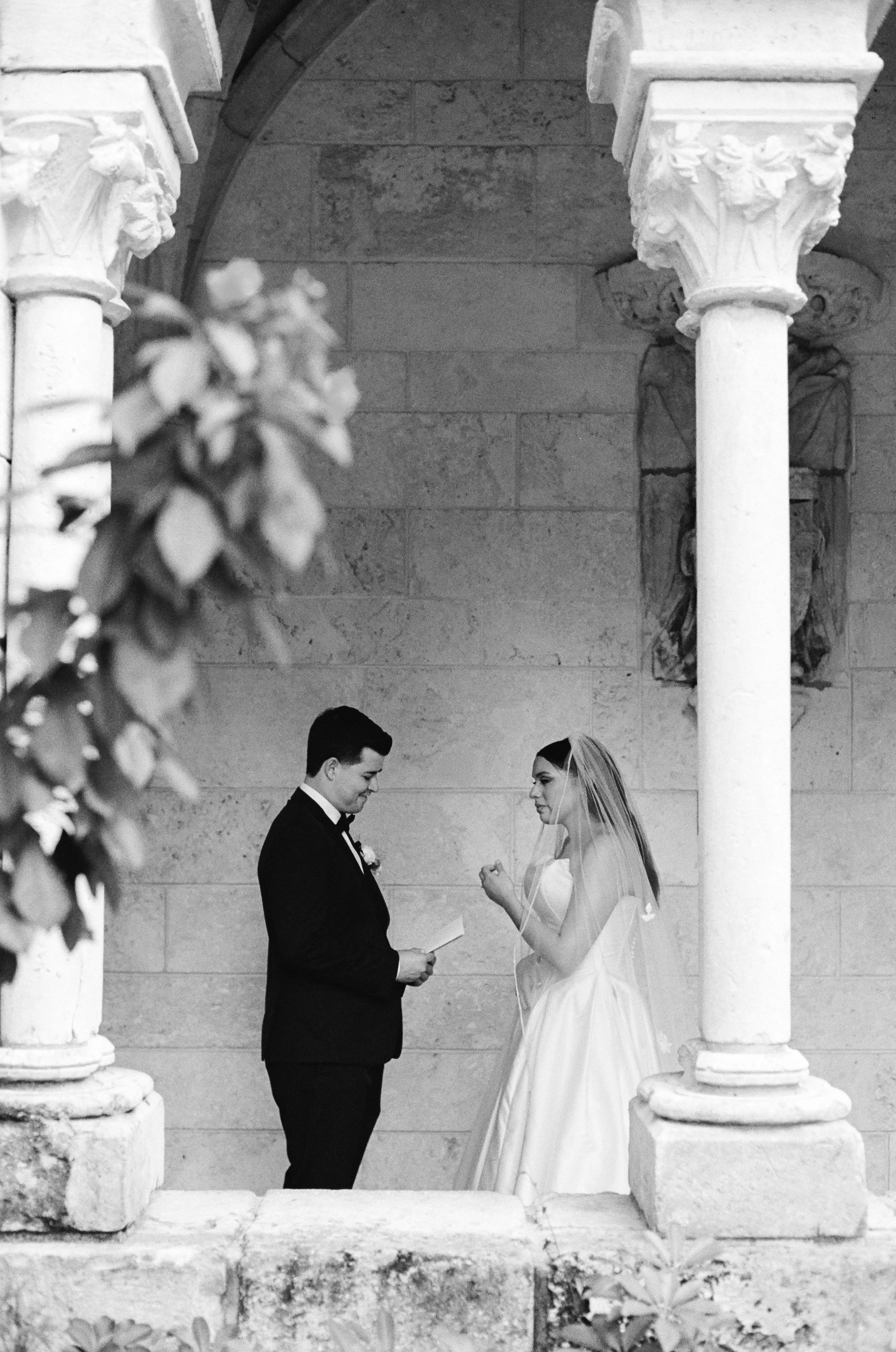 A black and white photo of a bride and groom standing in front of an ornate stone wall. The bride is wearing a wedding dress and veil, holding the groom's hands, who is in a tuxedo. They are seen through a decorative stone arch with floral embellishm