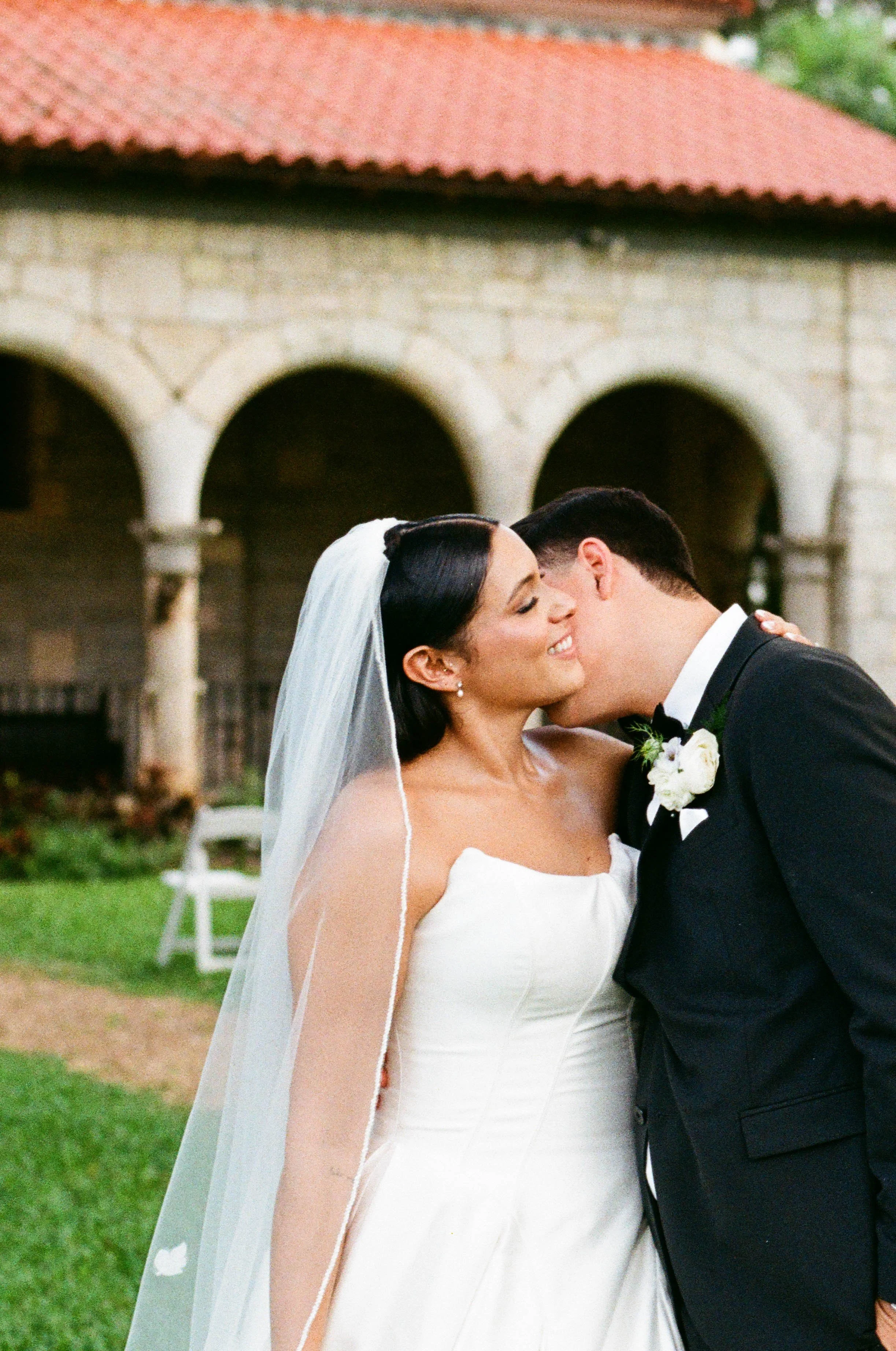 A bride and groom share a kiss on their wedding day outdoors, with greenery and a stone building with arches in the background.