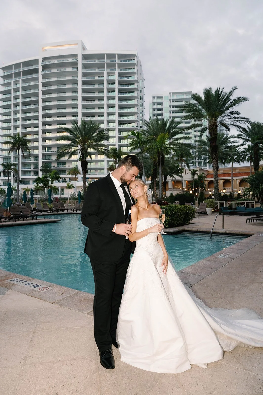 A bride and groom in wedding attire standing by a pool at a hotel or resort with high-rise buildings and palm trees in the background.