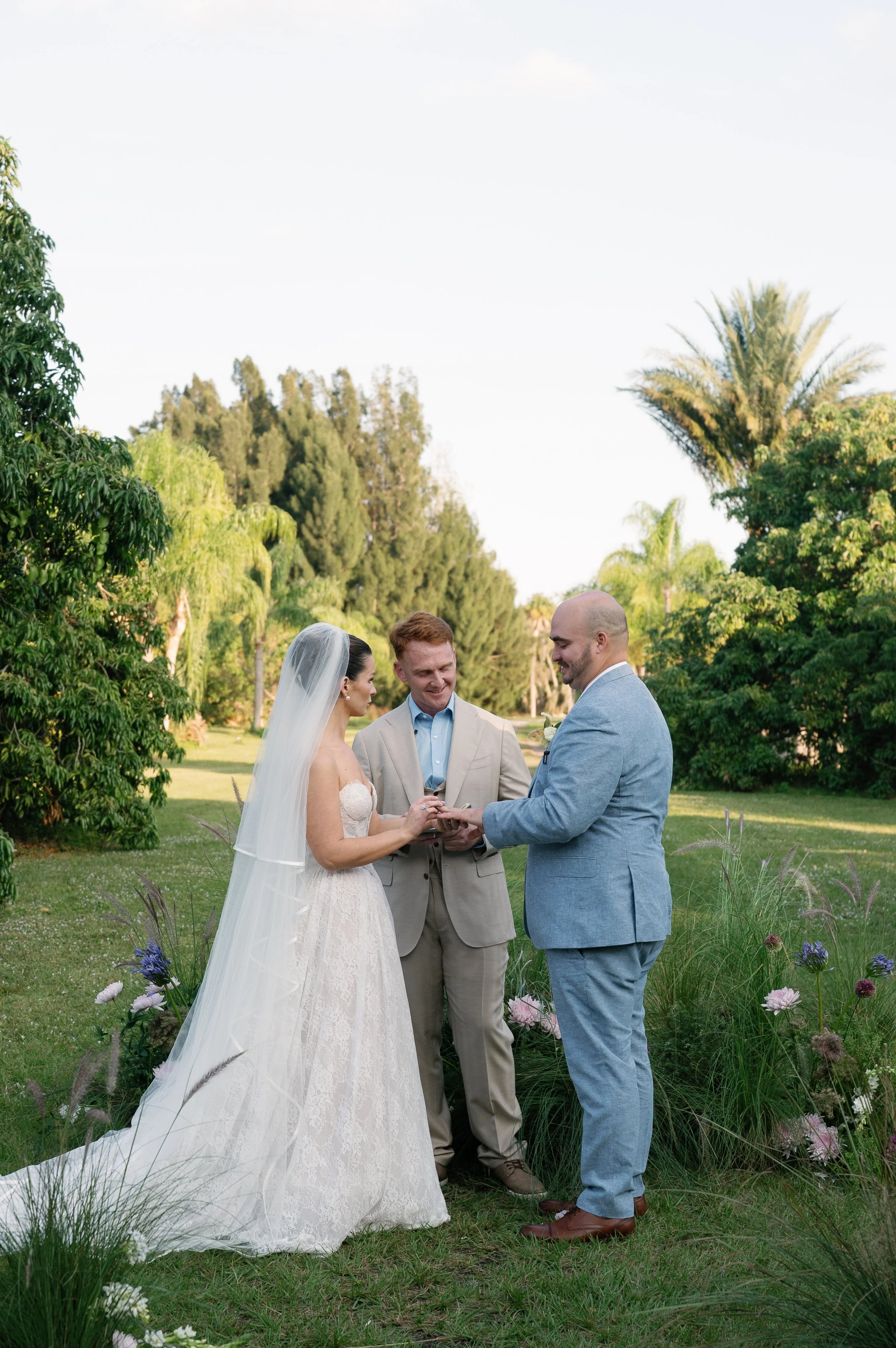 A wedding ceremony taking place outdoors, with a bride and groom exchanging vows, officiant standing between them, all surrounded by greenery and flowers. The bride is wearing a white dress with a veil, and the groom is dressed in a light gray suit.