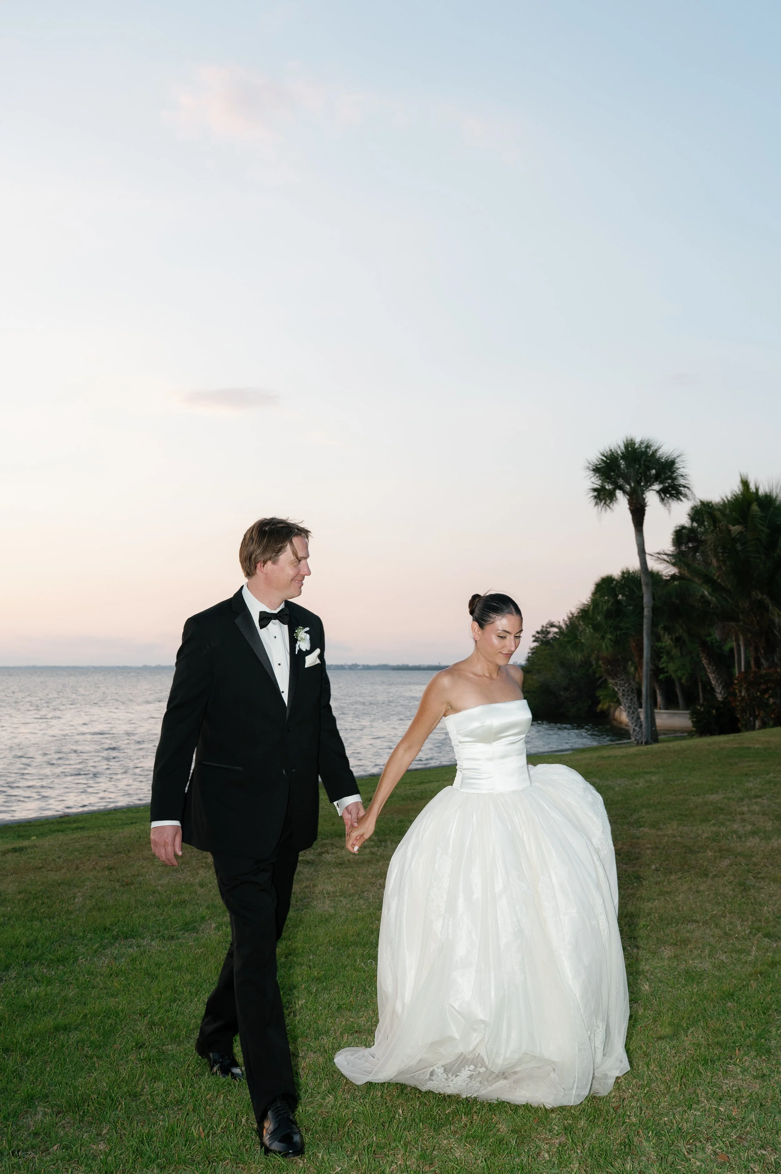 A bride and groom walking hand in hand on a grassy area near a body of water during sunset. The groom is wearing a black tuxedo with a bow tie, and the bride is wearing a strapless white wedding gown. There are palm trees and bushes in the background