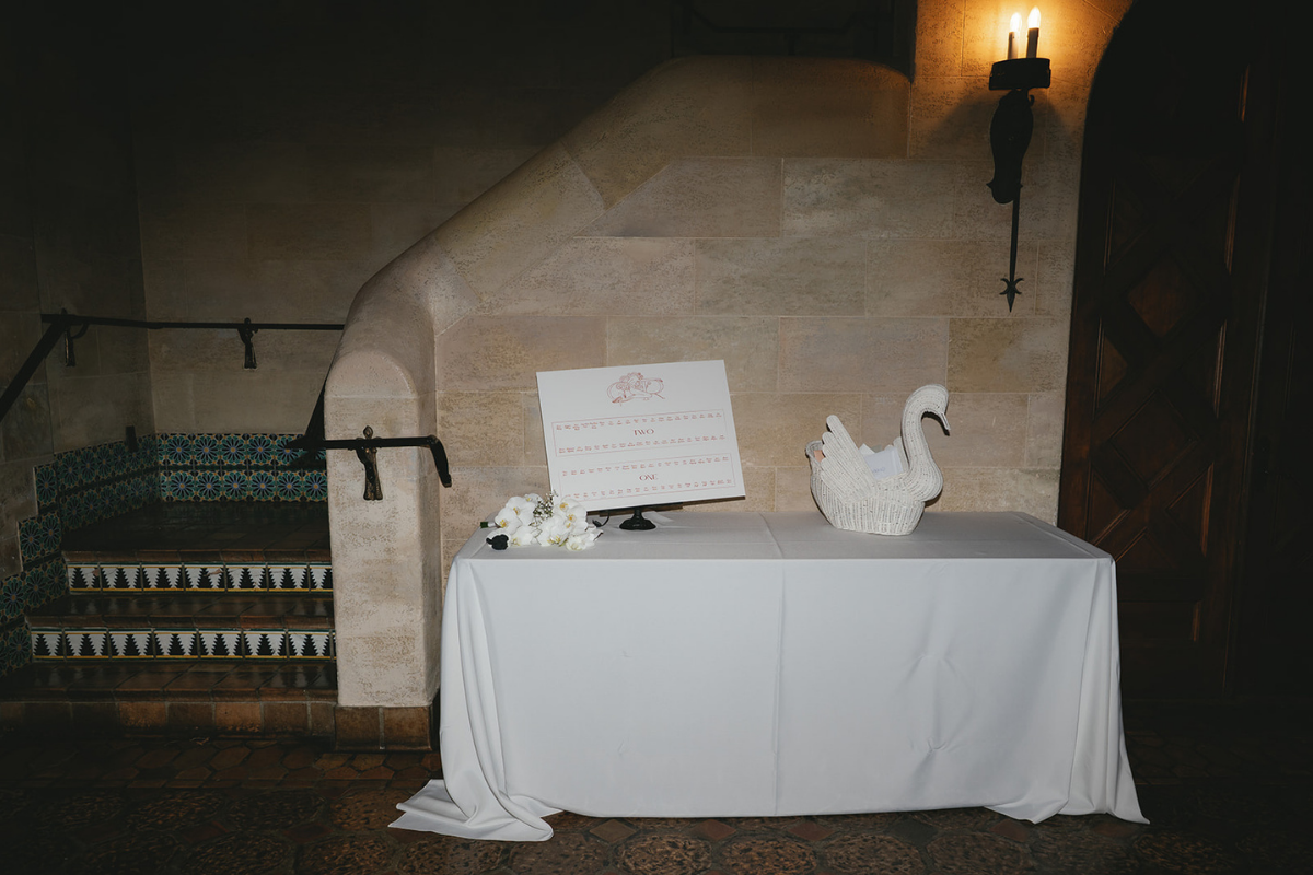 A white table with a folded white tablecloth in a dimly lit room, featuring a white swan-shaped basket, a small stand with a sign, and some flowers, against a stone wall and a staircase with colorful tile risers.