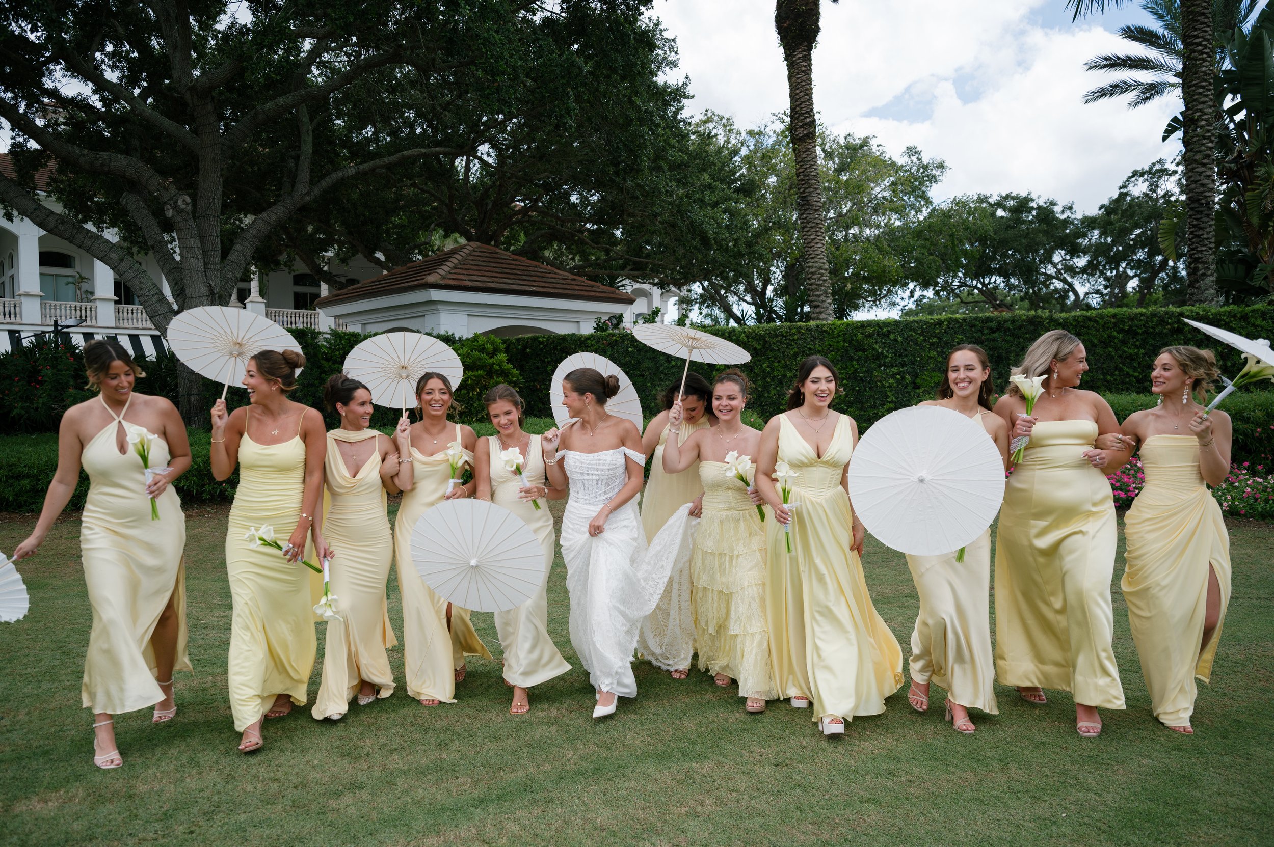 A group of women in yellow dresses holding white parasols and white calla lilies, walking outdoors on green grass with trees and a white building in the background.