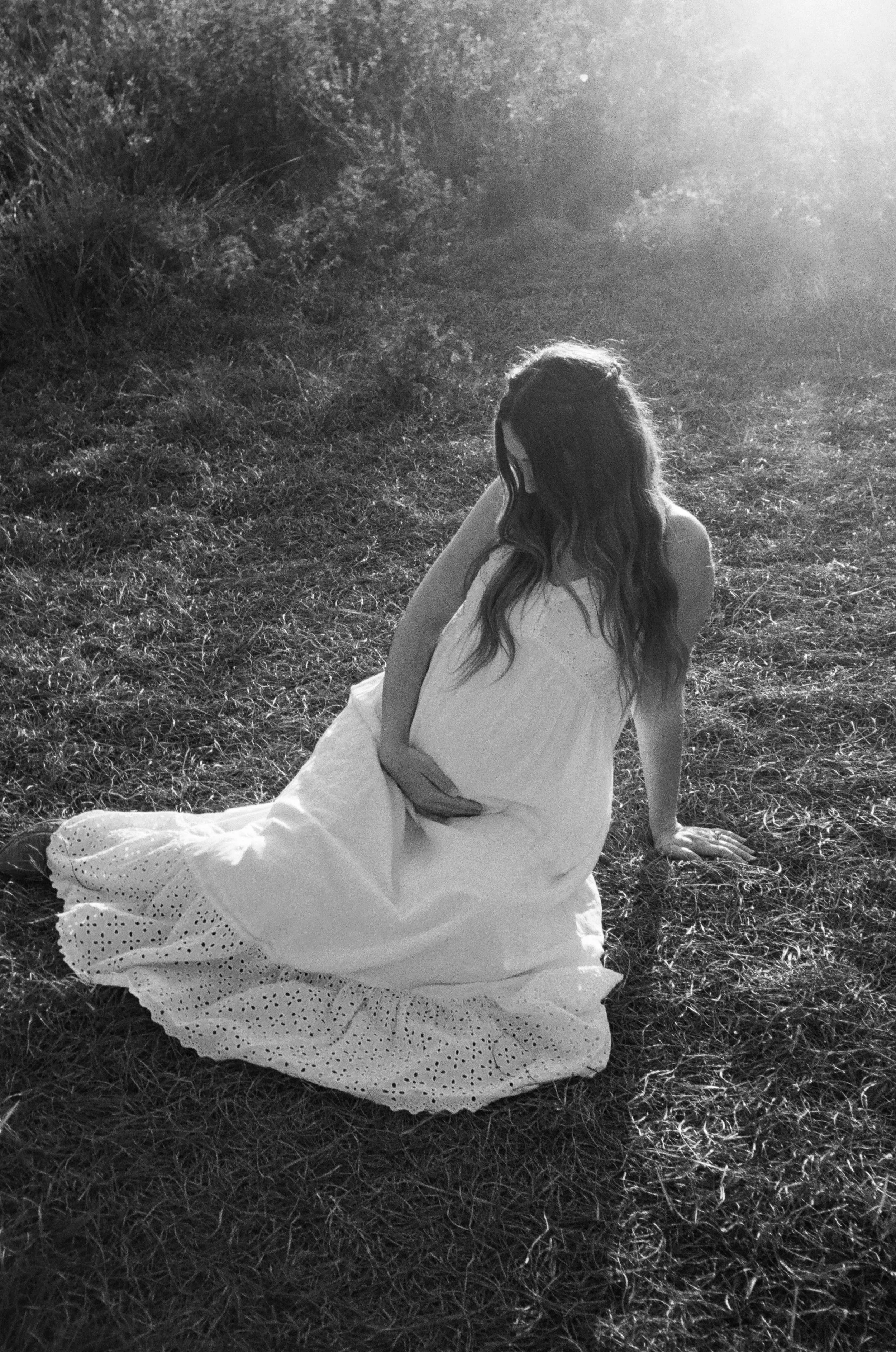 A woman with long wavy hair wearing a white dress with eyelet details, sitting on the grass with her legs extended and her left arm supporting her, in a natural outdoor setting with grass and bushes, illuminated by sunlight.