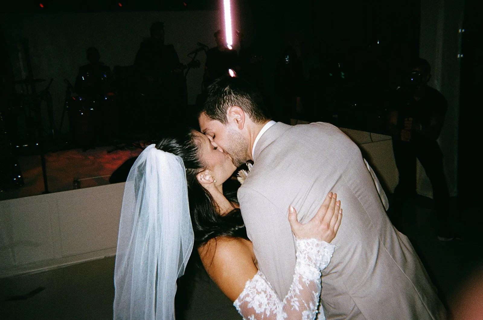 A bride and groom share a kiss during their wedding celebration with guests in the background.