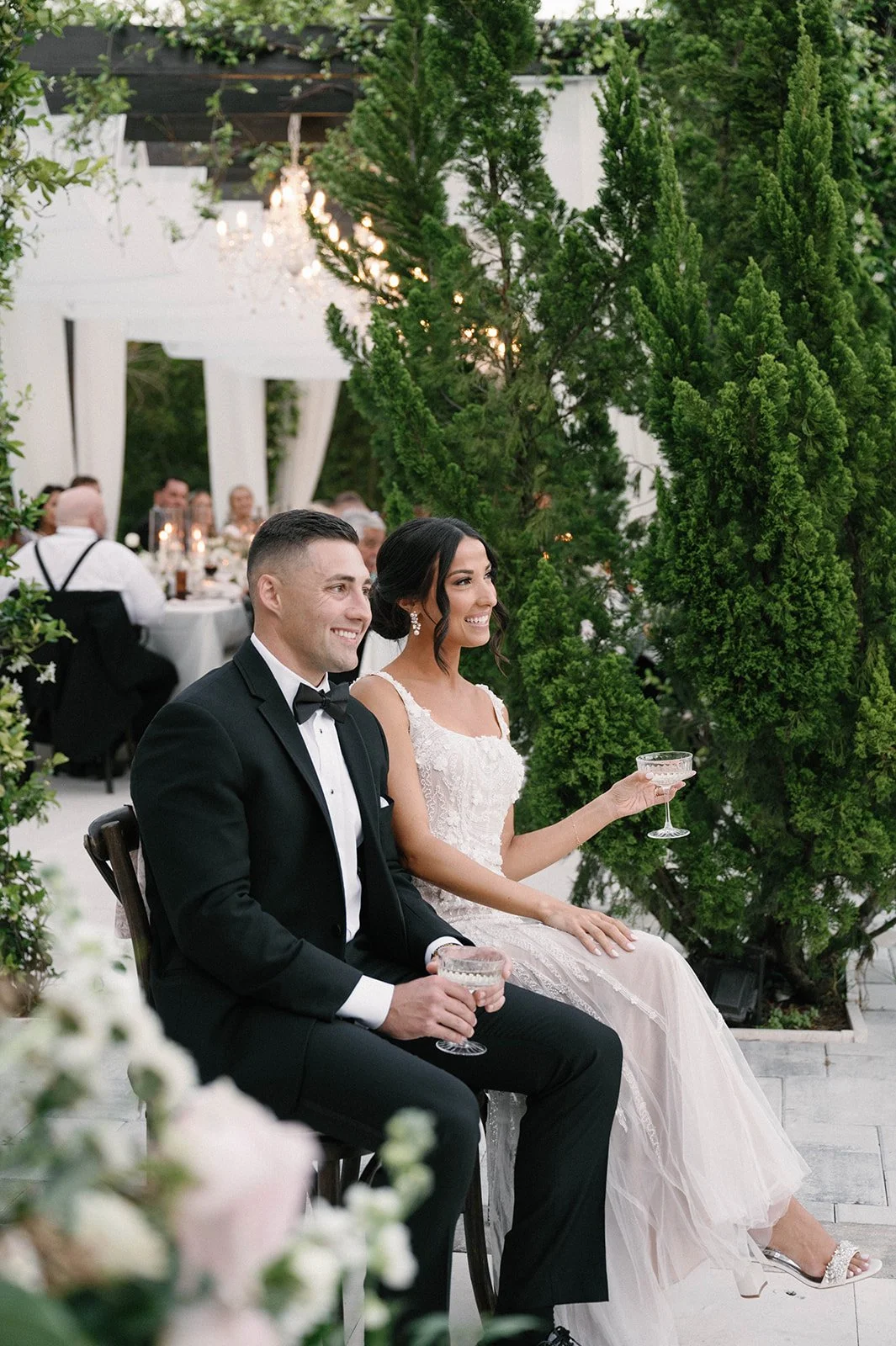 A bride and groom sitting together at their wedding reception, smiling and holding glasses, with greenery and trees in the background.