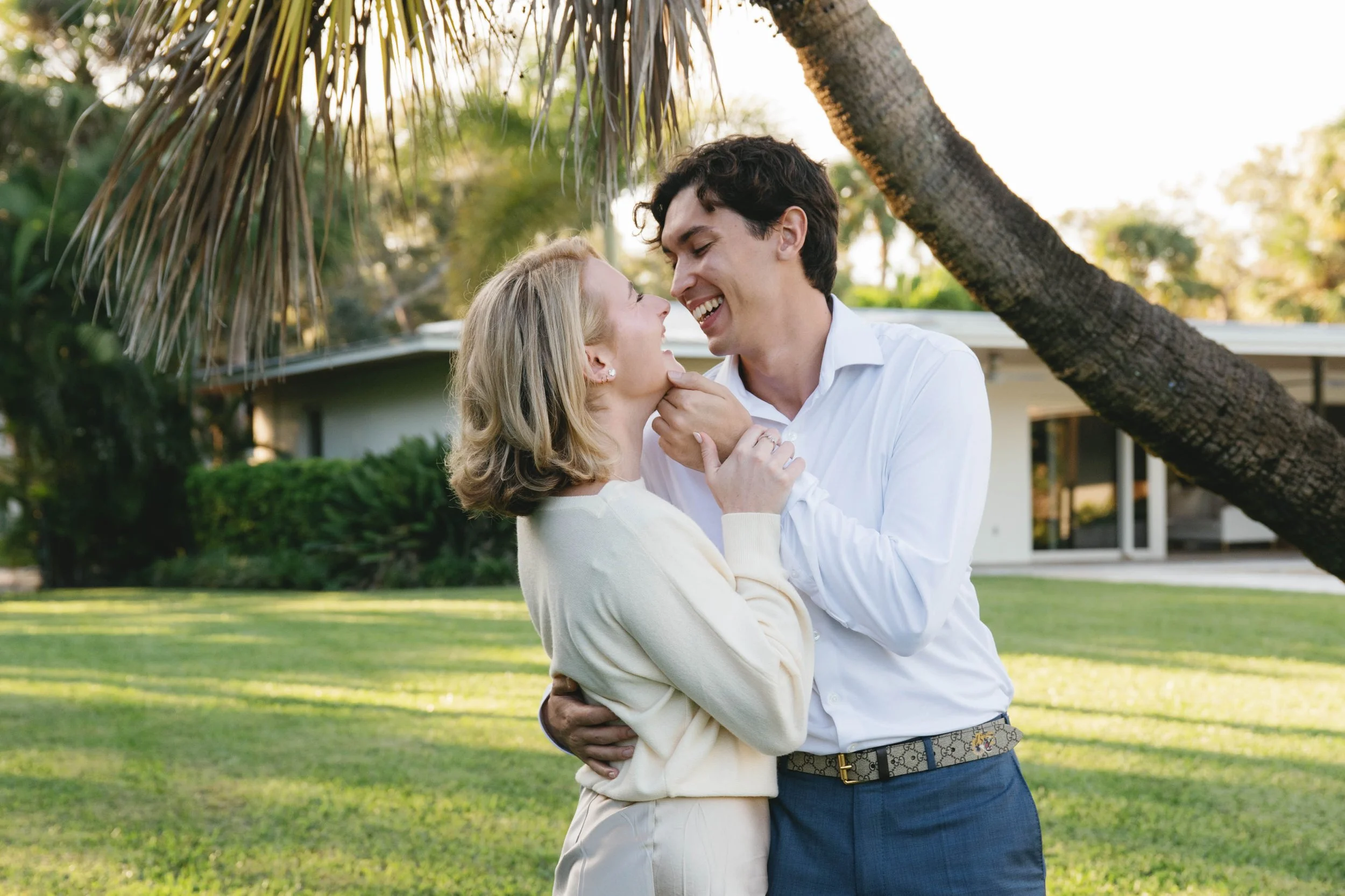 A man and woman embracing outdoors beneath a tree, smiling and looking into each other's eyes.