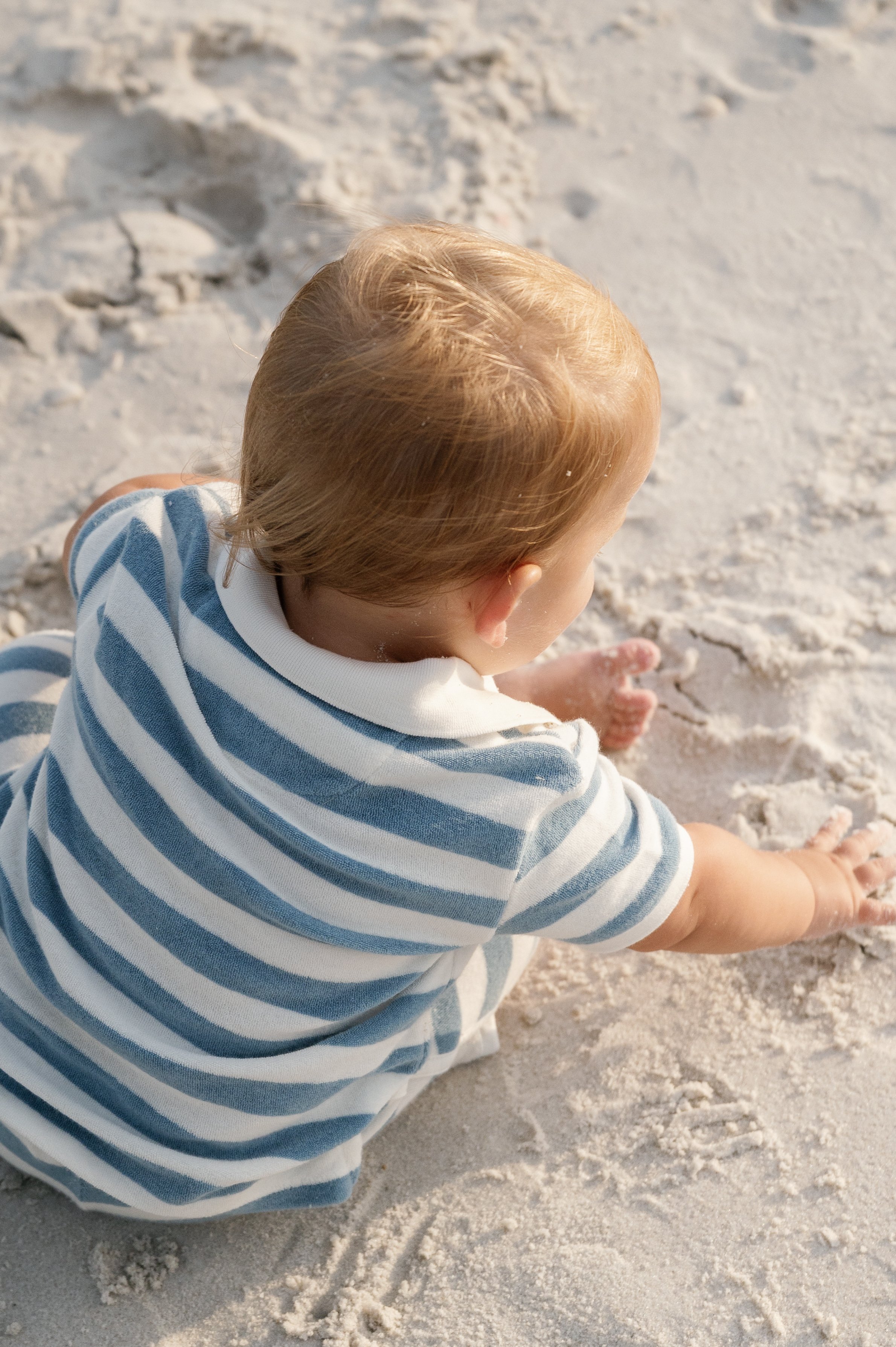 A young child with blonde hair and light skin sitting on sandy beach, wearing a white and blue striped shirt, and playing with the sand.