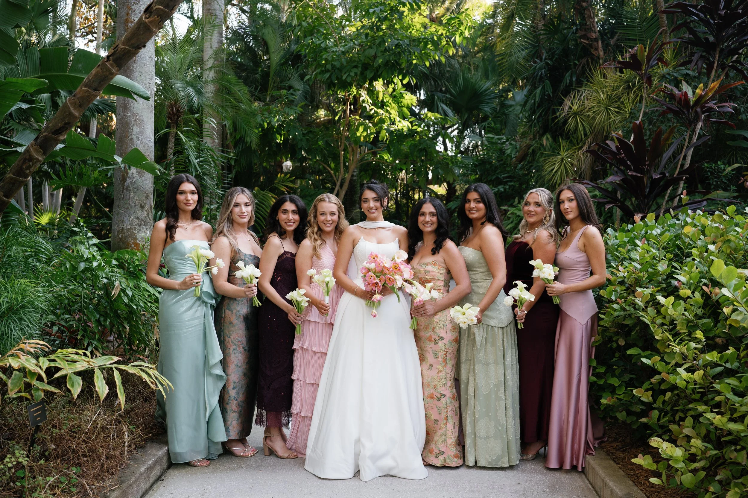 A group of women in elegant dresses standing together in a lush, green outdoor garden setting, holding flowers and smiling.