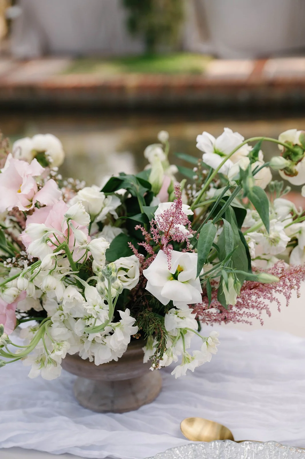 A floral arrangement with pink and white flowers and greenery in a stone bowl, placed on a white textured surface with a gold spoon nearby, outdoors with blurry background of a garden and stone fountain.