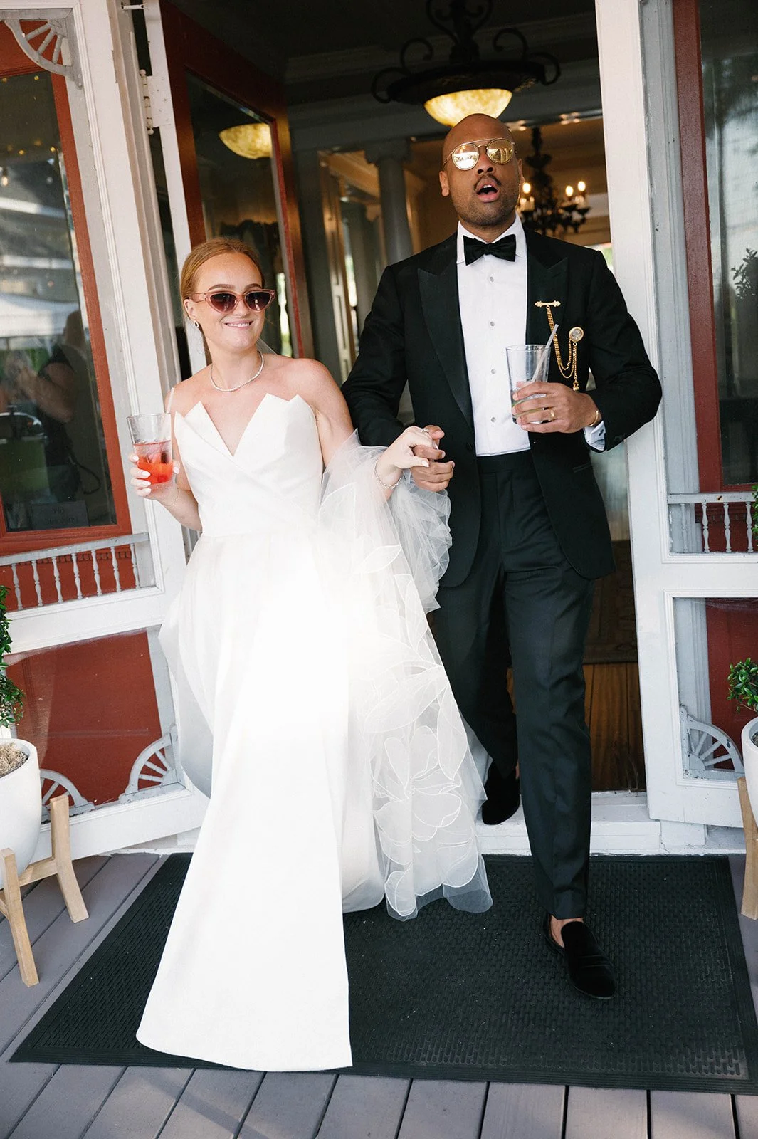 A bride and groom walking out of a house, holding hands. The bride wears a white wedding dress and sunglasses, holding a drink. The groom is dressed in a tuxedo with a bow tie, wearing sunglasses, and holding a glass.