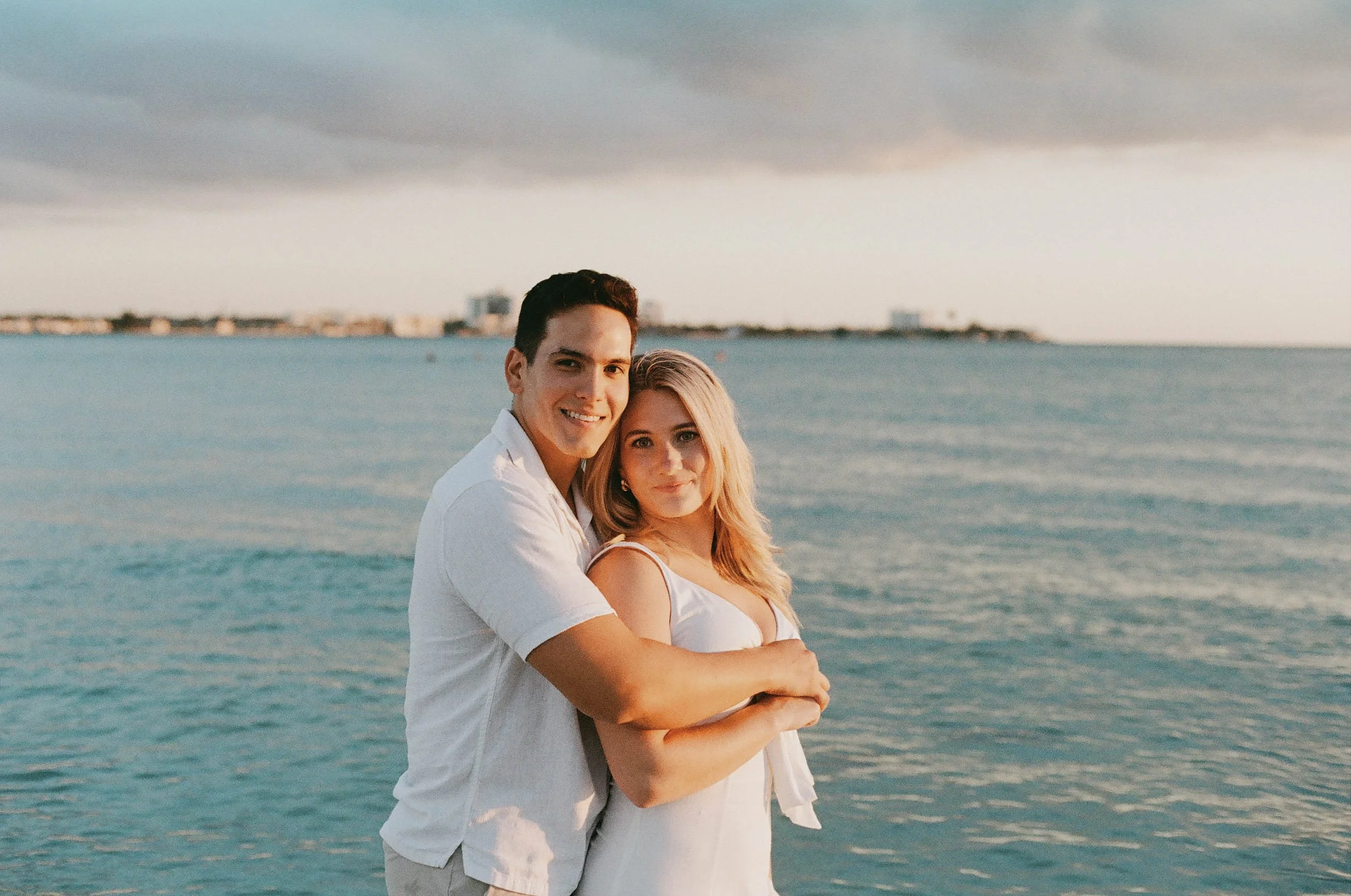 A young couple standing by the water at a beach during sunset, smiling and embracing.
