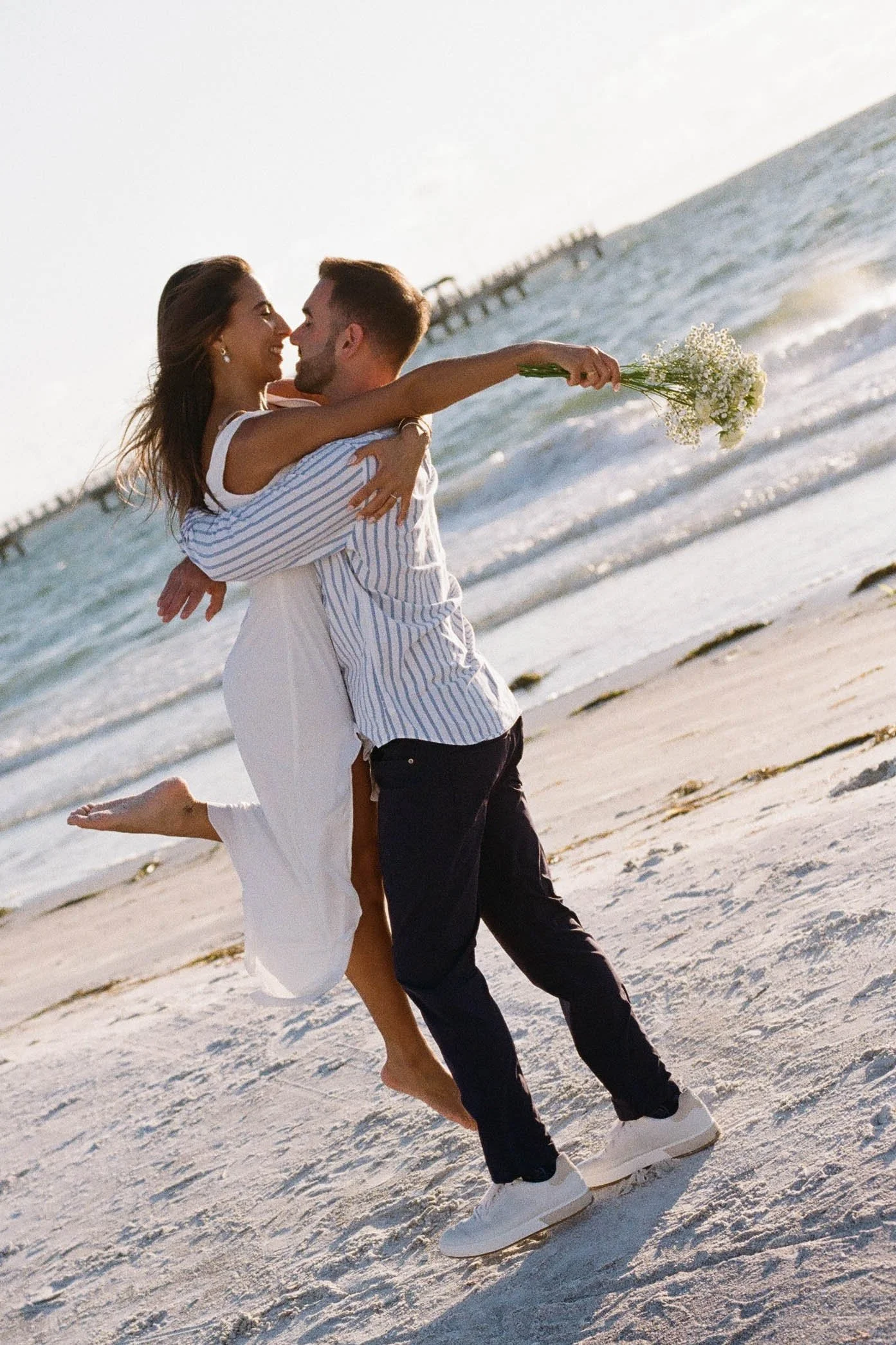 A couple is on the beach, with one person holding a bouquet of flowers, and they are about to kiss. They are standing on the sand near the ocean with a pier in the background.
