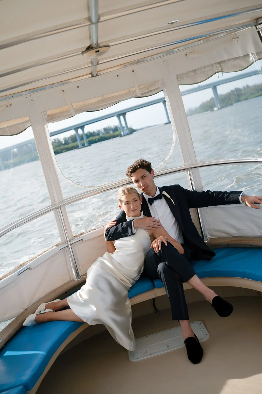 A young couple in formal attire sitting together on a boat, with water and a bridge in the background.