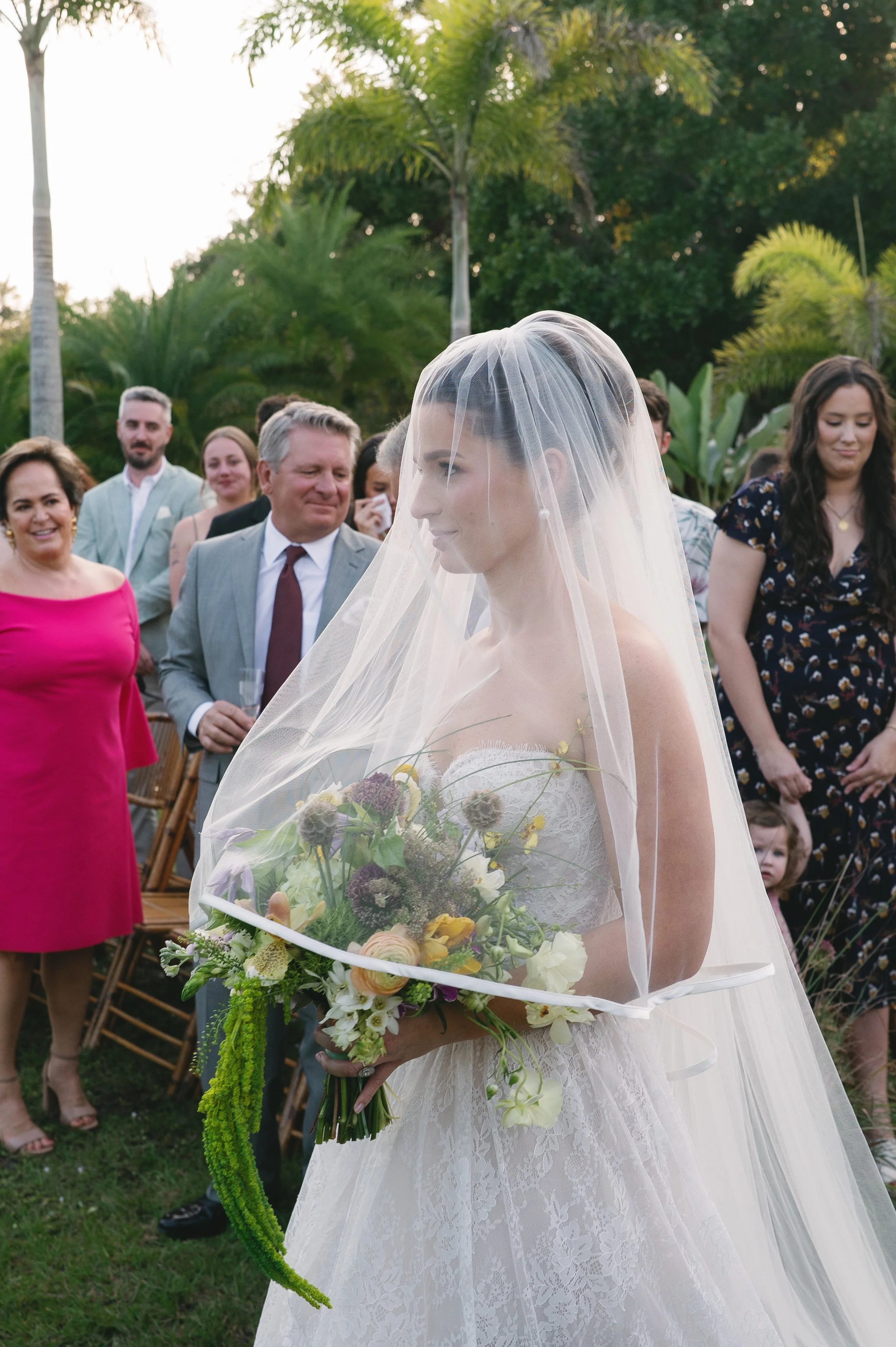 A bride in a white wedding dress and veil holding a bouquet of flowers at an outdoor wedding ceremony. Guests are smiling and watching her in a lush garden setting with tall trees and green foliage.