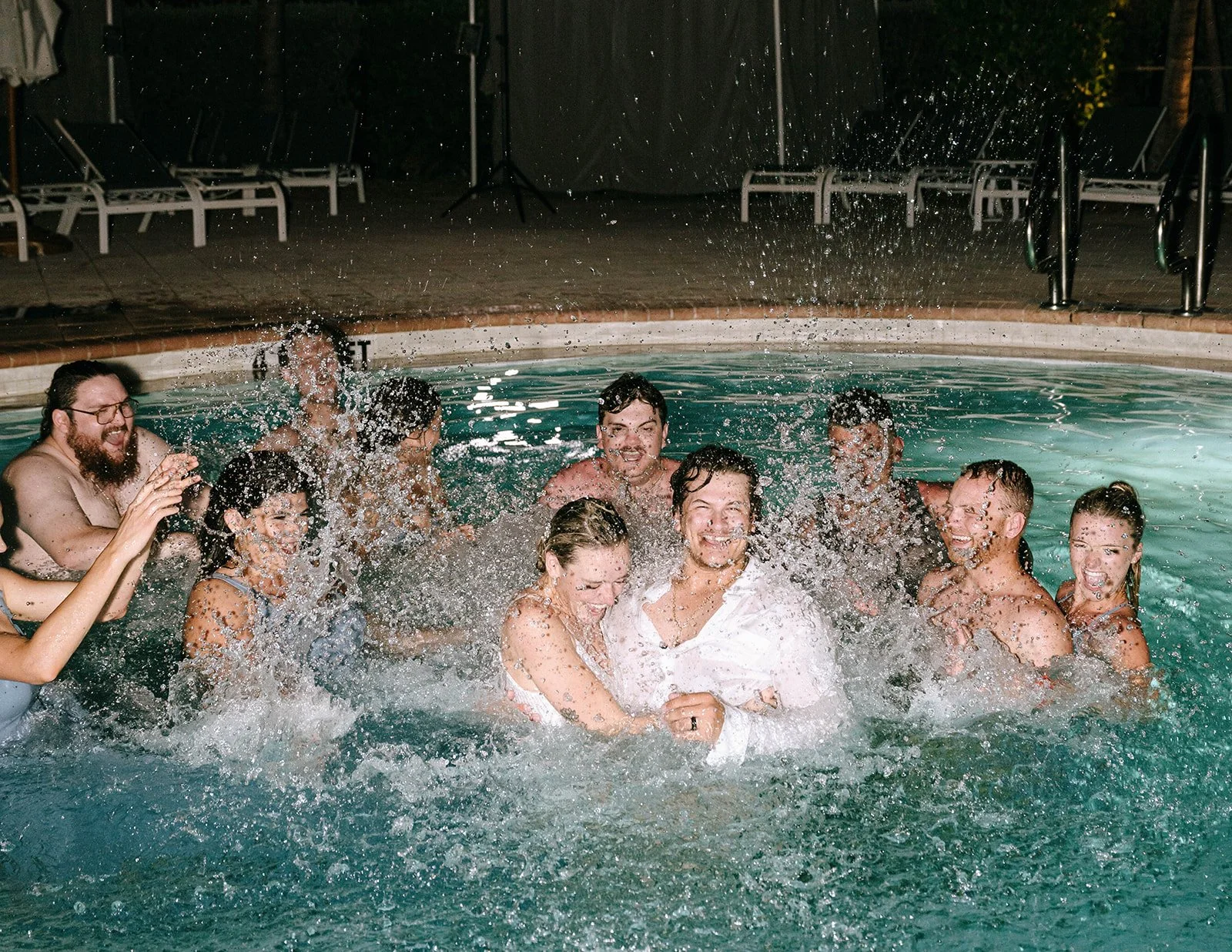 A group of people enjoying a joyful moment in a swimming pool at night, splashing water and smiling.