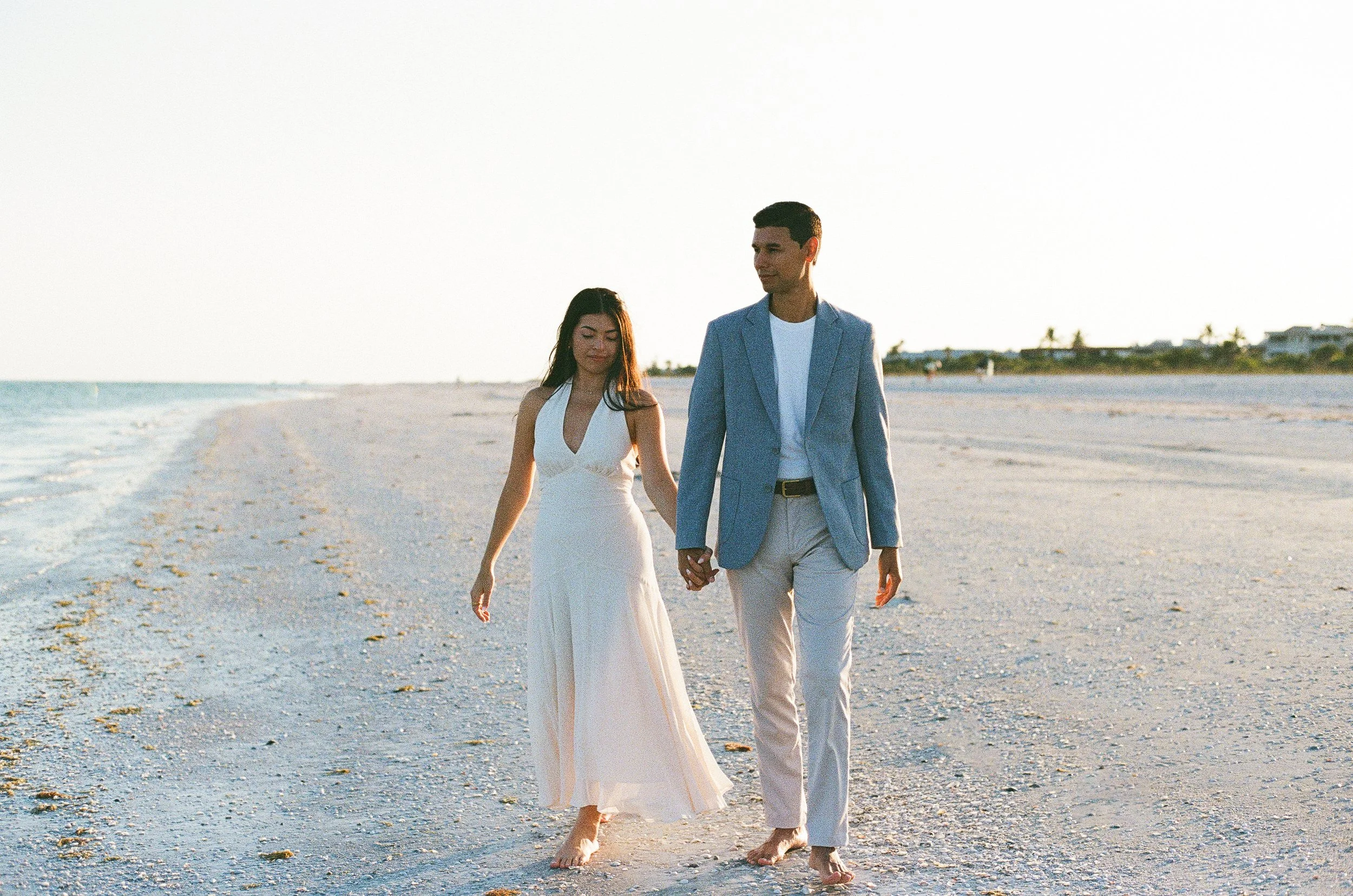 A couple holding hands walking barefoot on a sandy beach near the ocean during sunset.