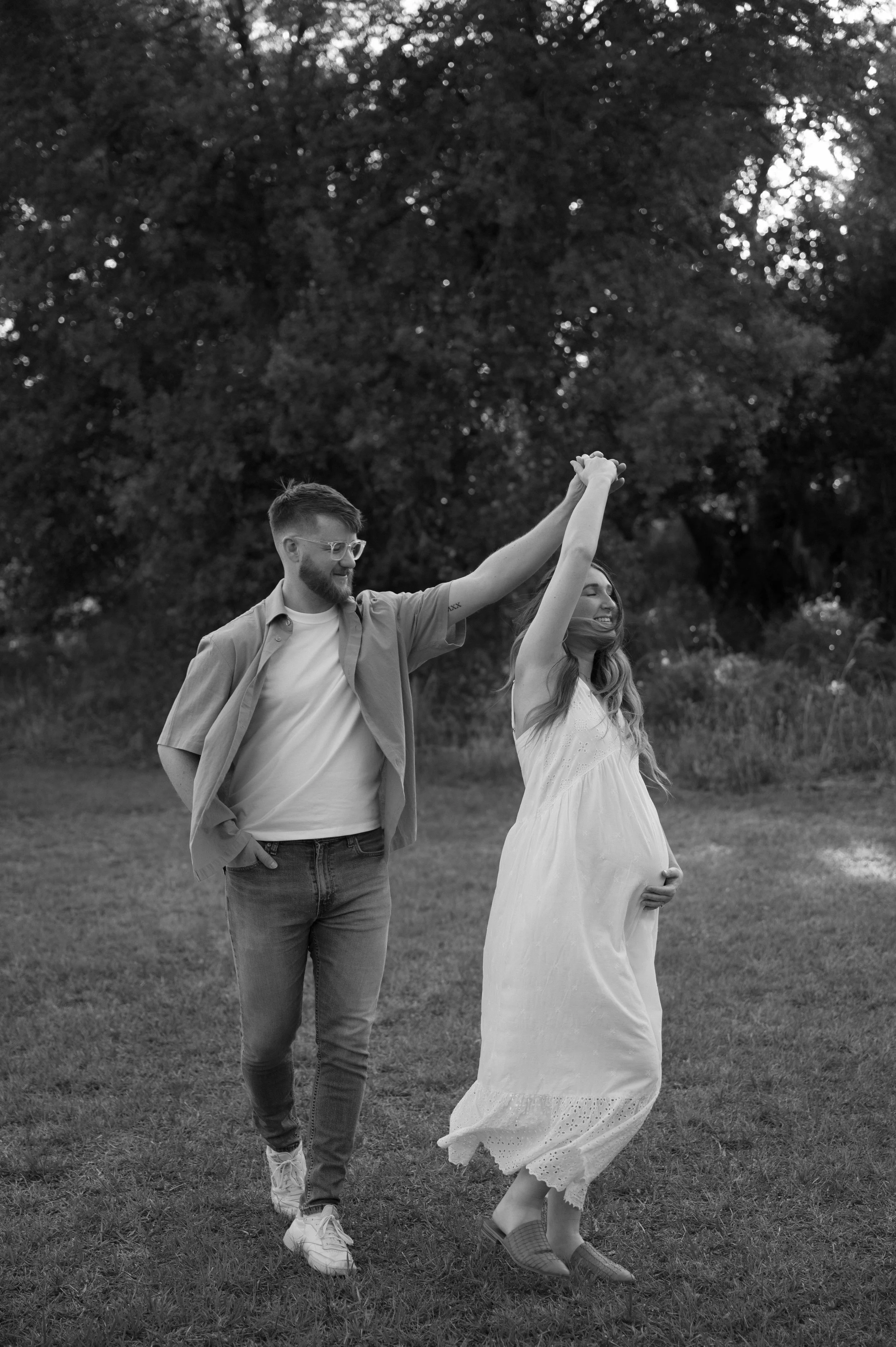 A couple dancing outdoors in a grassy area with trees in the background. The man has a beard, wears glasses, a light shirt over a t-shirt, and jeans. The woman wears a flowy dress and is smiling with her eyes closed. They hold hands and are smiling.