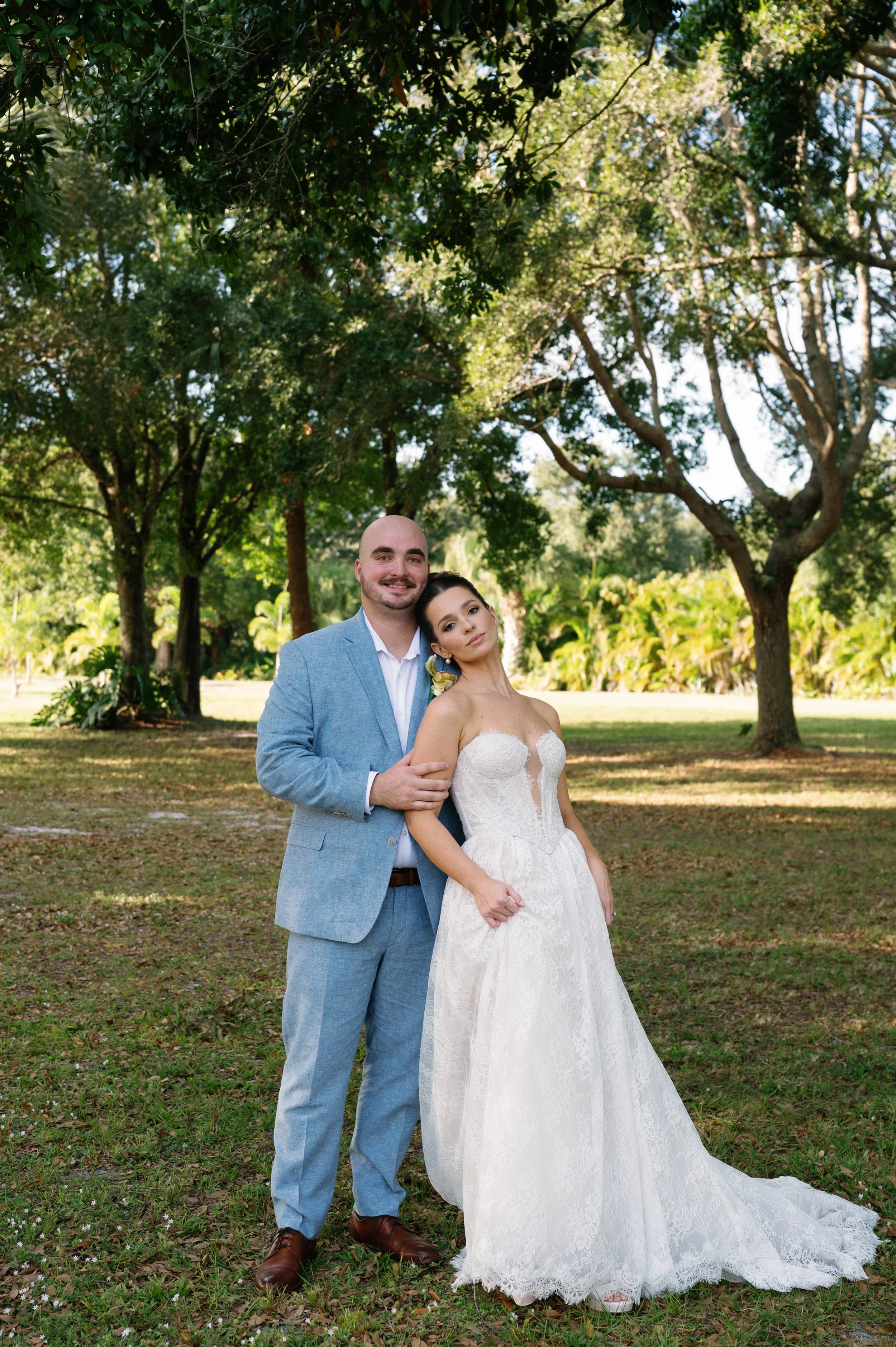 A bride and groom standing together outdoors on grass with trees in the background. The groom is wearing a light blue suit and brown shoes, while the bride is wearing a strapless lace wedding gown with a train and has a flower in her hair. They are s